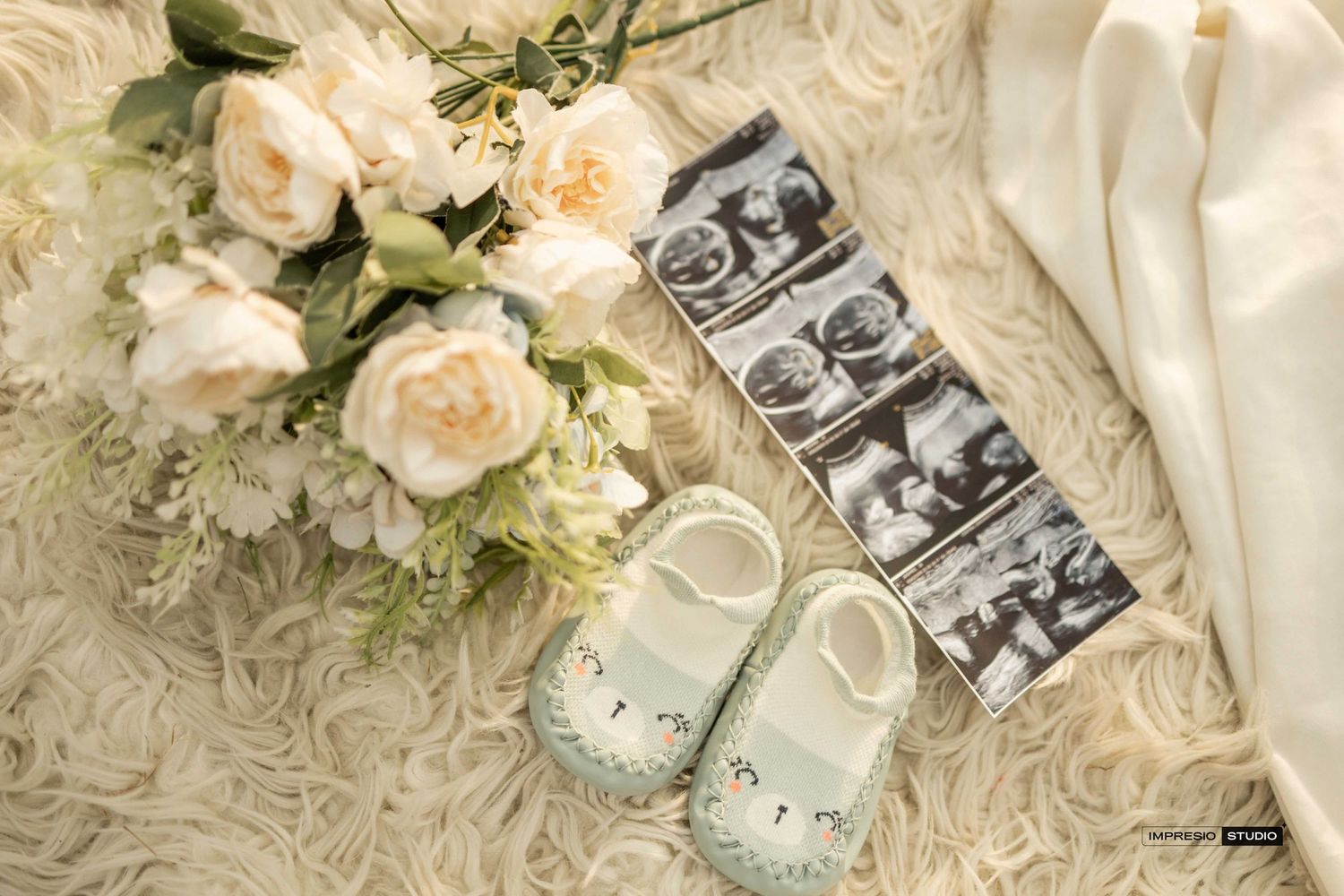 Baby shoes, ultrasound photos, and white roses bouquet arranged on a soft white fur surface for a pregnancy announcement.