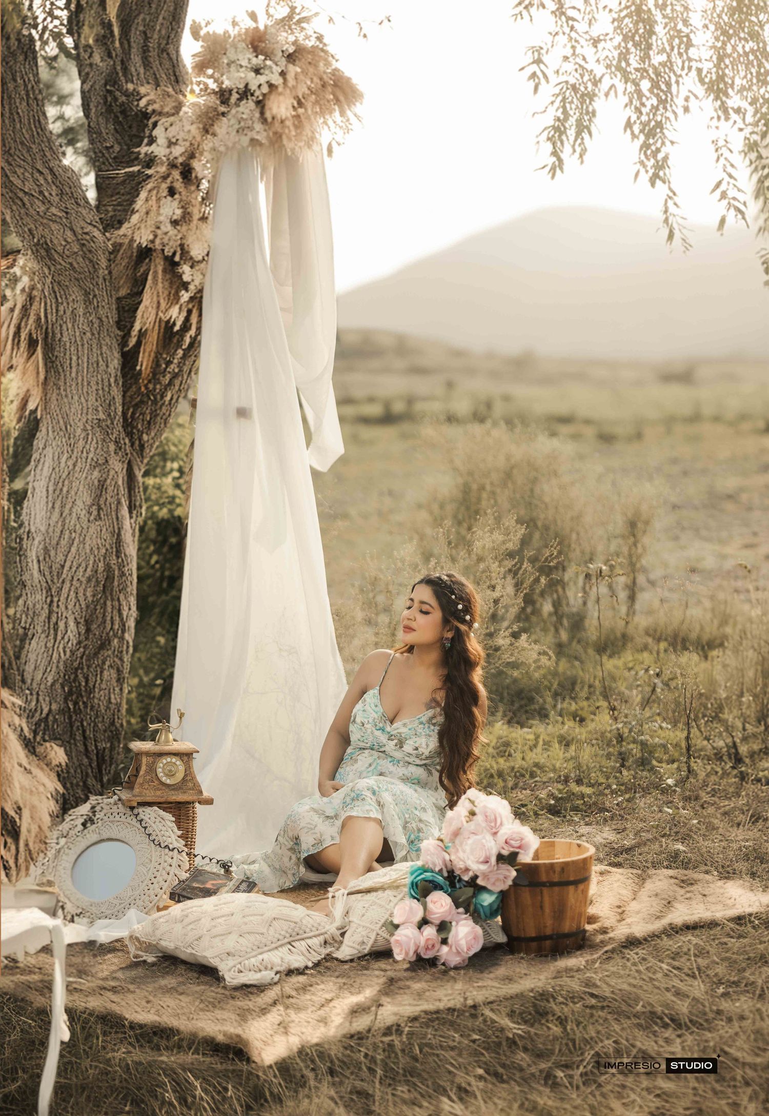 A woman in a floral dress sits on a blanket outdoors near a hanging white gown, flowers, and vintage decor in a scenic field.