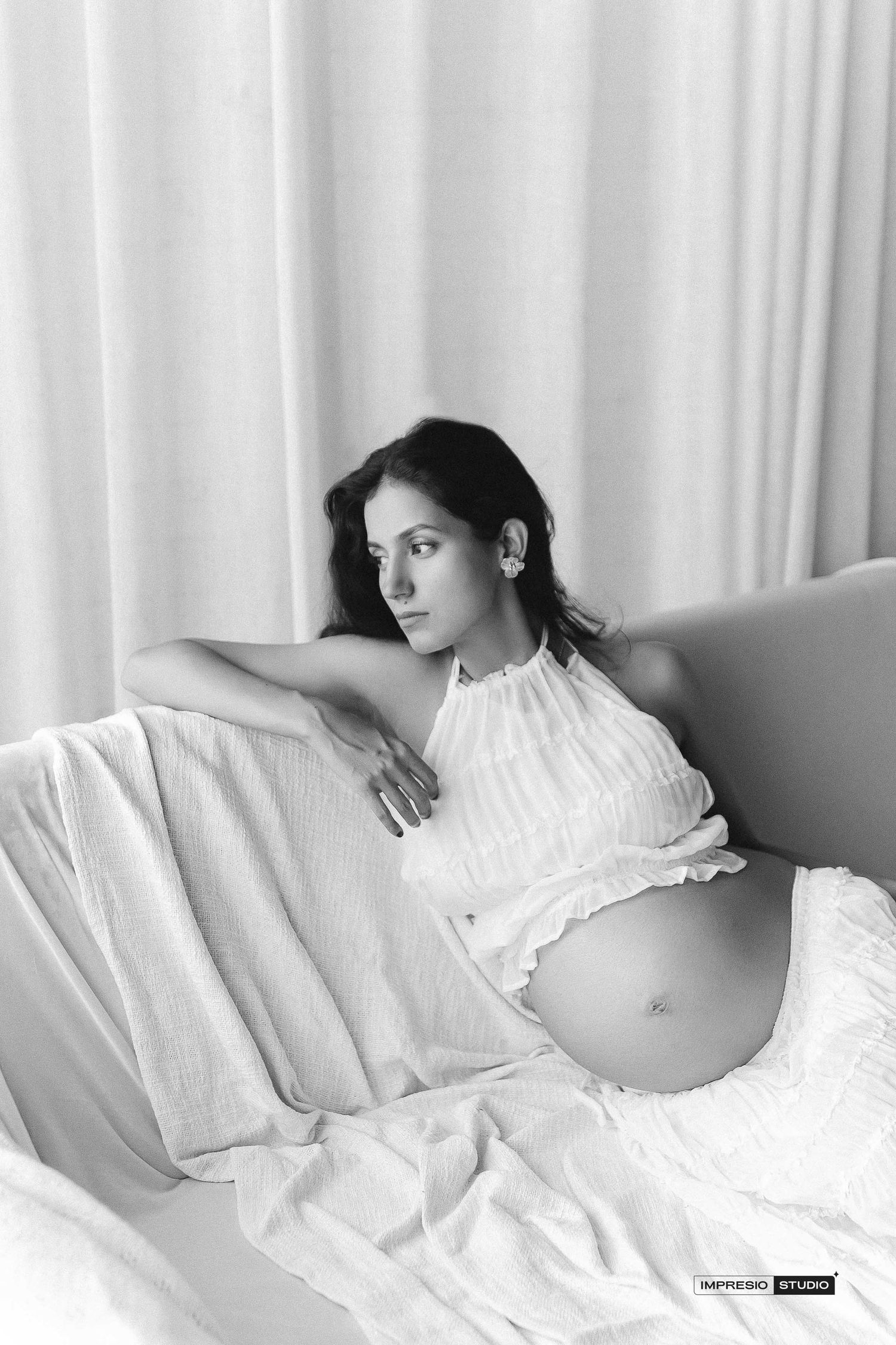 Black and white maternity photo of a pregnant woman in a white halter dress, reclining gracefully on a bed.
