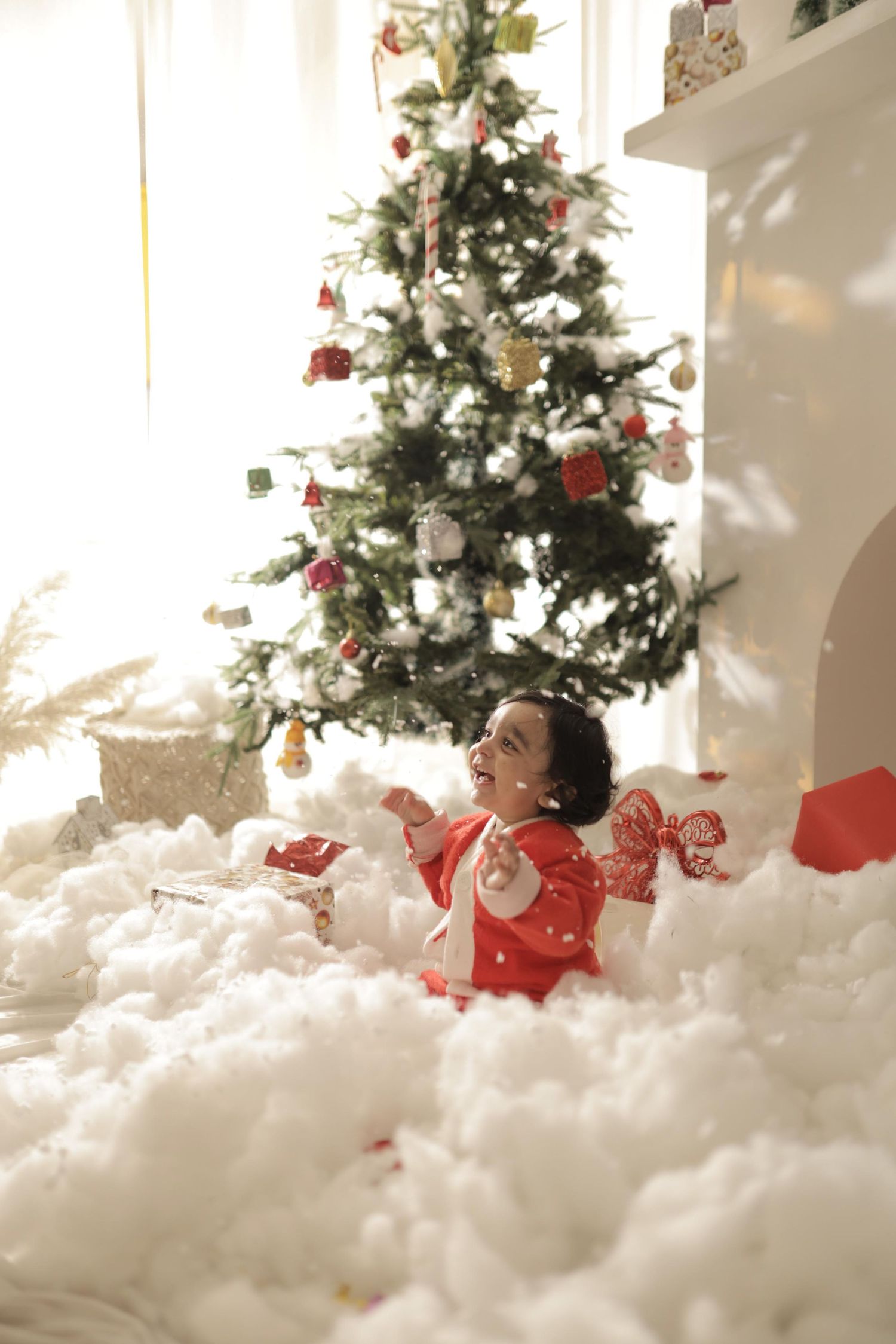 A joyful child in red pajamas plays in artificial snow beside a decorated Christmas tree near a fireplace.