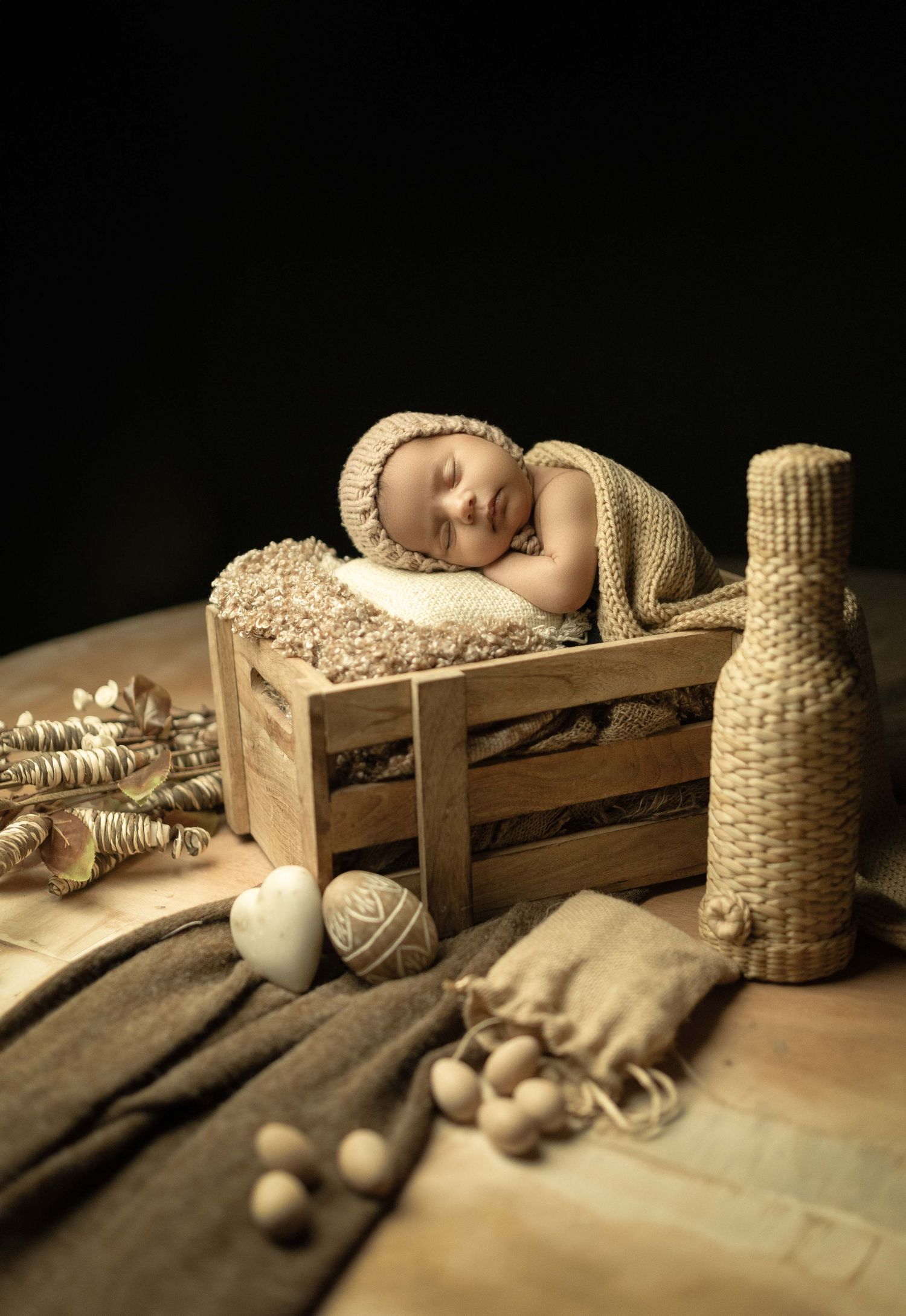 Sleeping newborn baby in knit hat resting in rustic wooden crate with burlap bottle and decorative props.