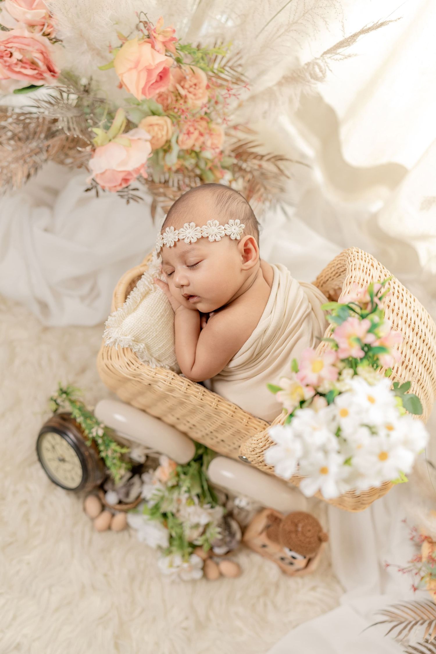 Sleeping newborn baby wrapped in cream swaddle with floral headband resting in wicker basket surrounded by flowers.
