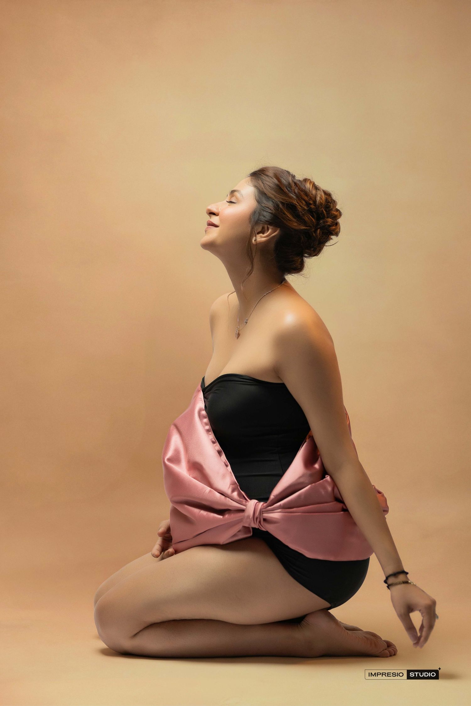 Woman in black strapless bodysuit with pink satin bow sits gracefully on floor, smiling with eyes closed.