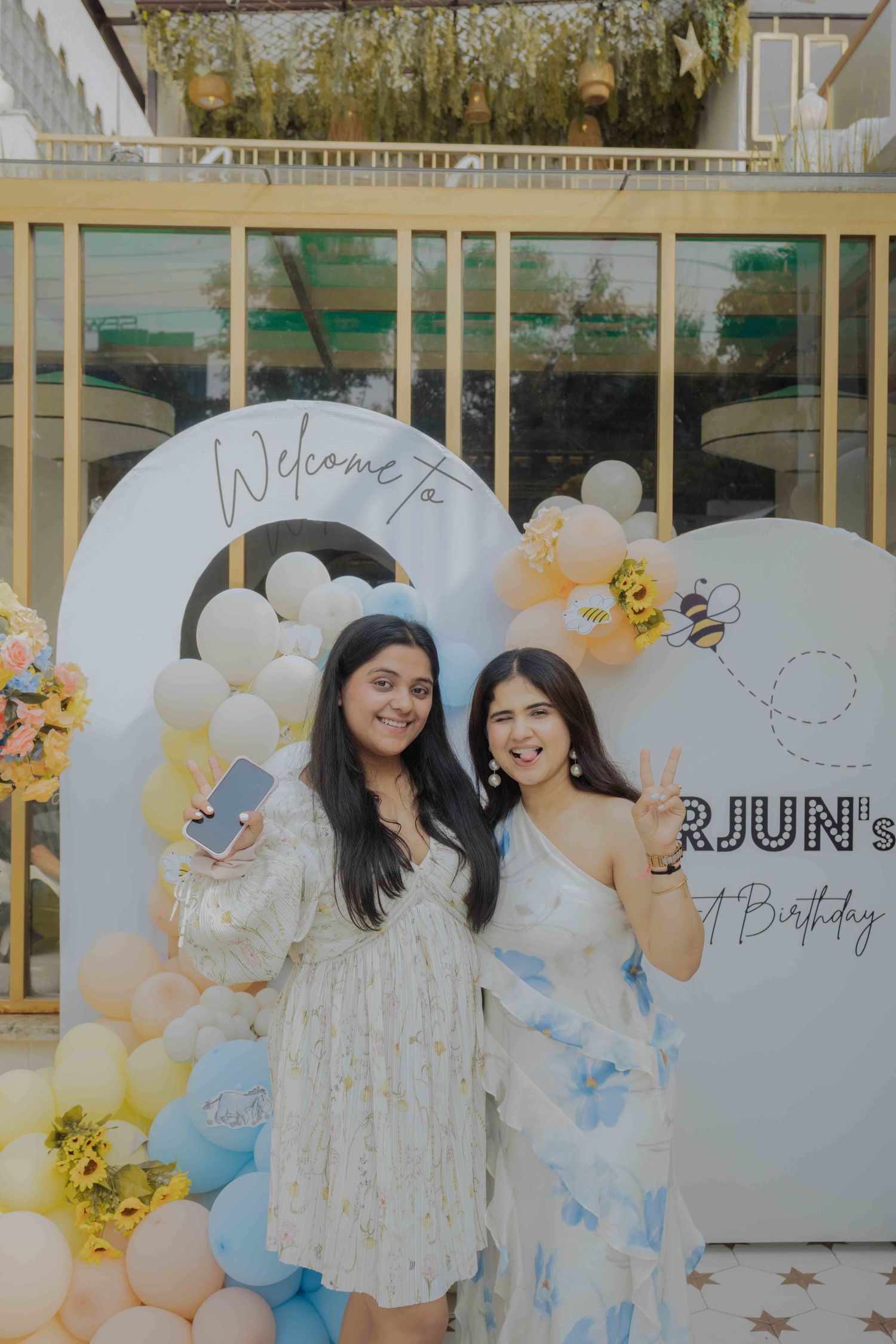 Two women smiling at a birthday party with balloon decorations and a welcome sign backdrop.