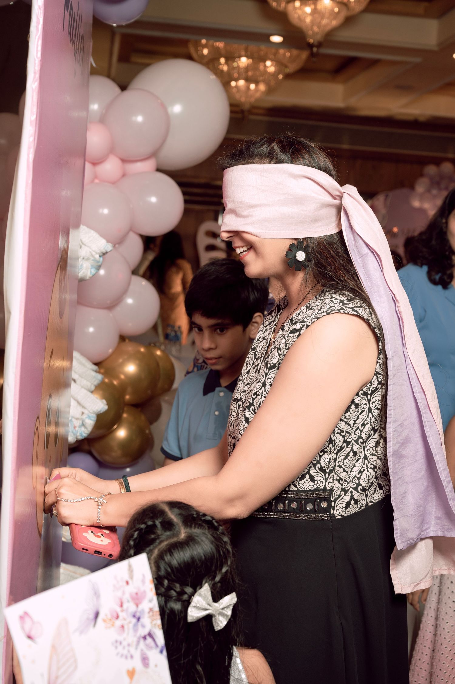 Blindfolded woman playing party game at elegant balloon-decorated celebration event with guests watching.
