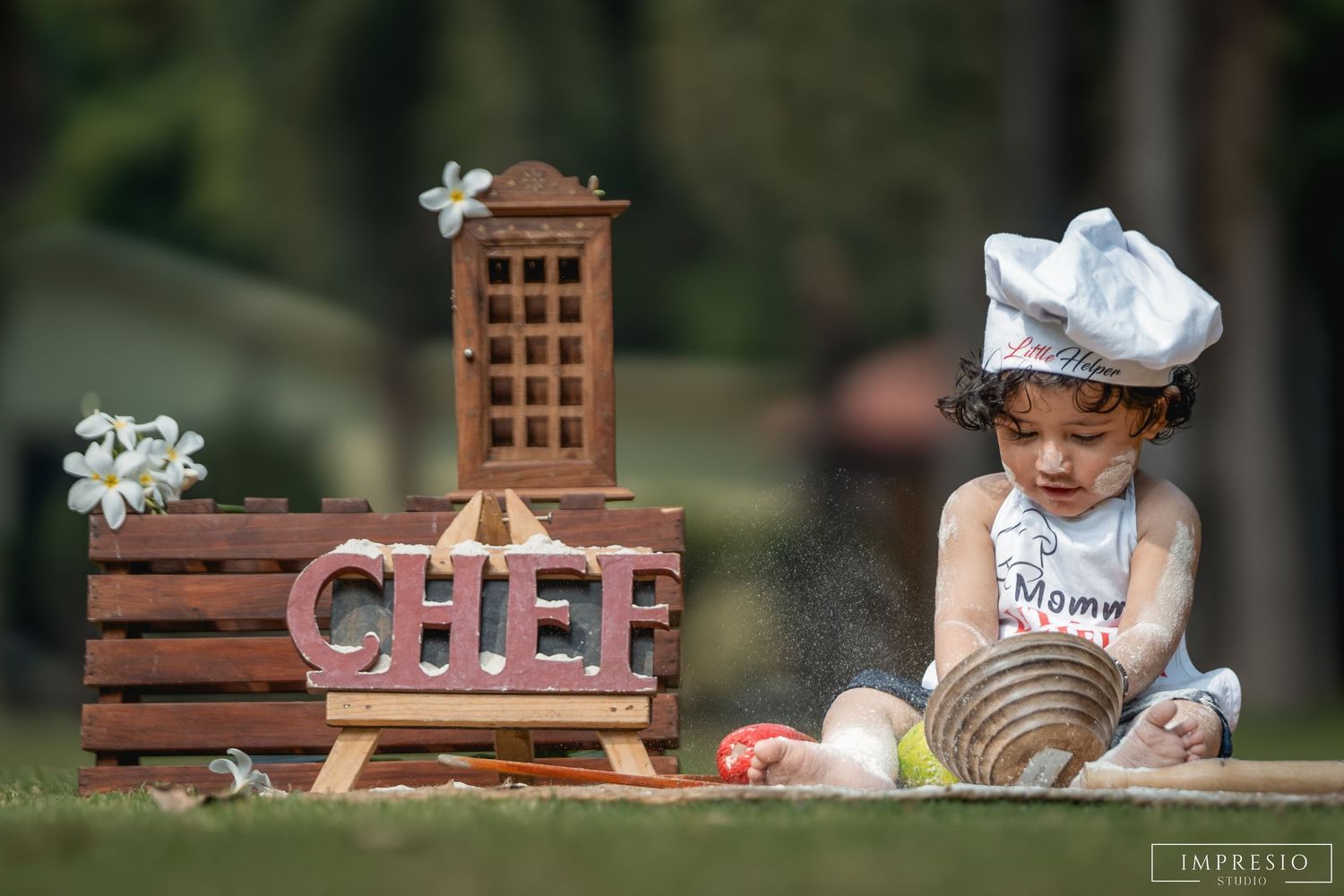 Adorable baby dressed as a chef in white hat and apron plays outdoors near a wooden CHEF sign with flowers.