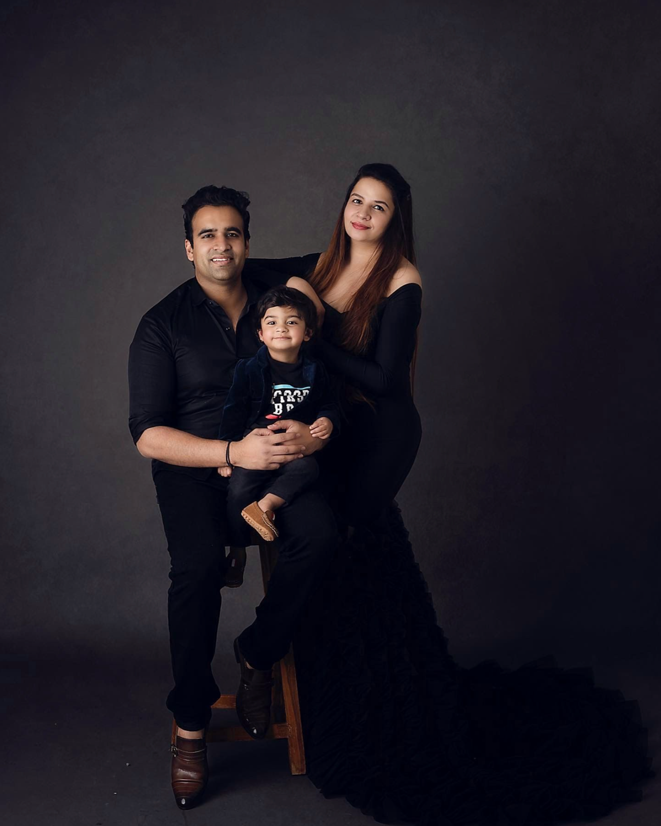 Family maternity portraits with couple and toddler in black outfits against dark backdrop.