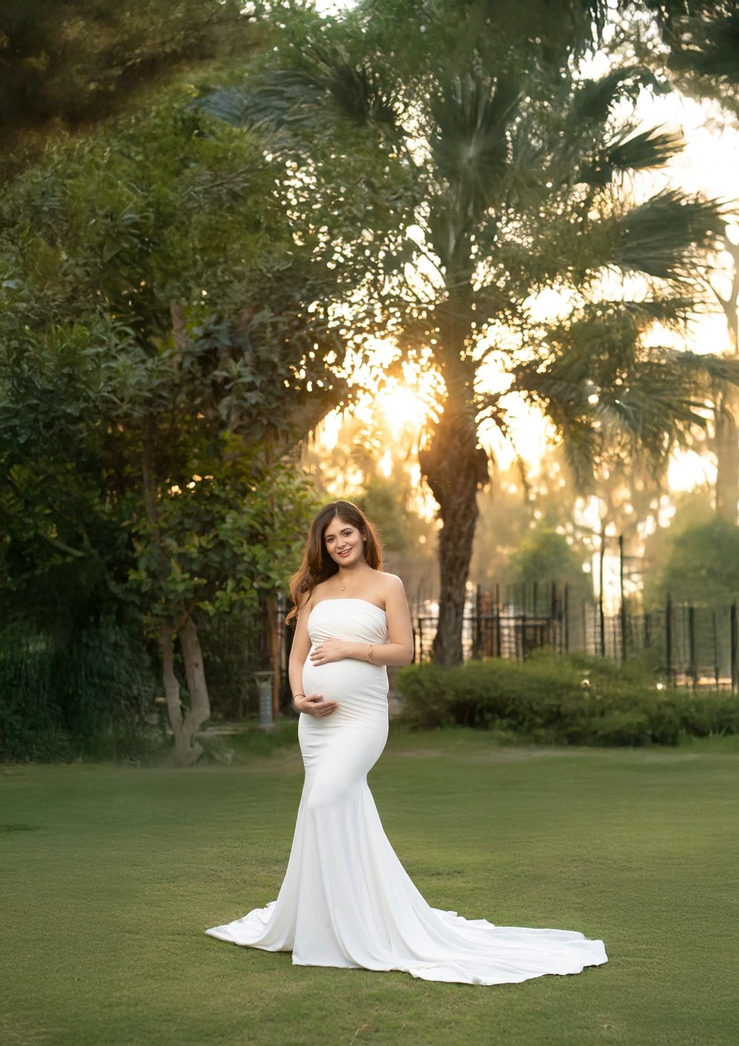 Golden hour maternity portrait in white fitted gown with palm trees and garden setting.