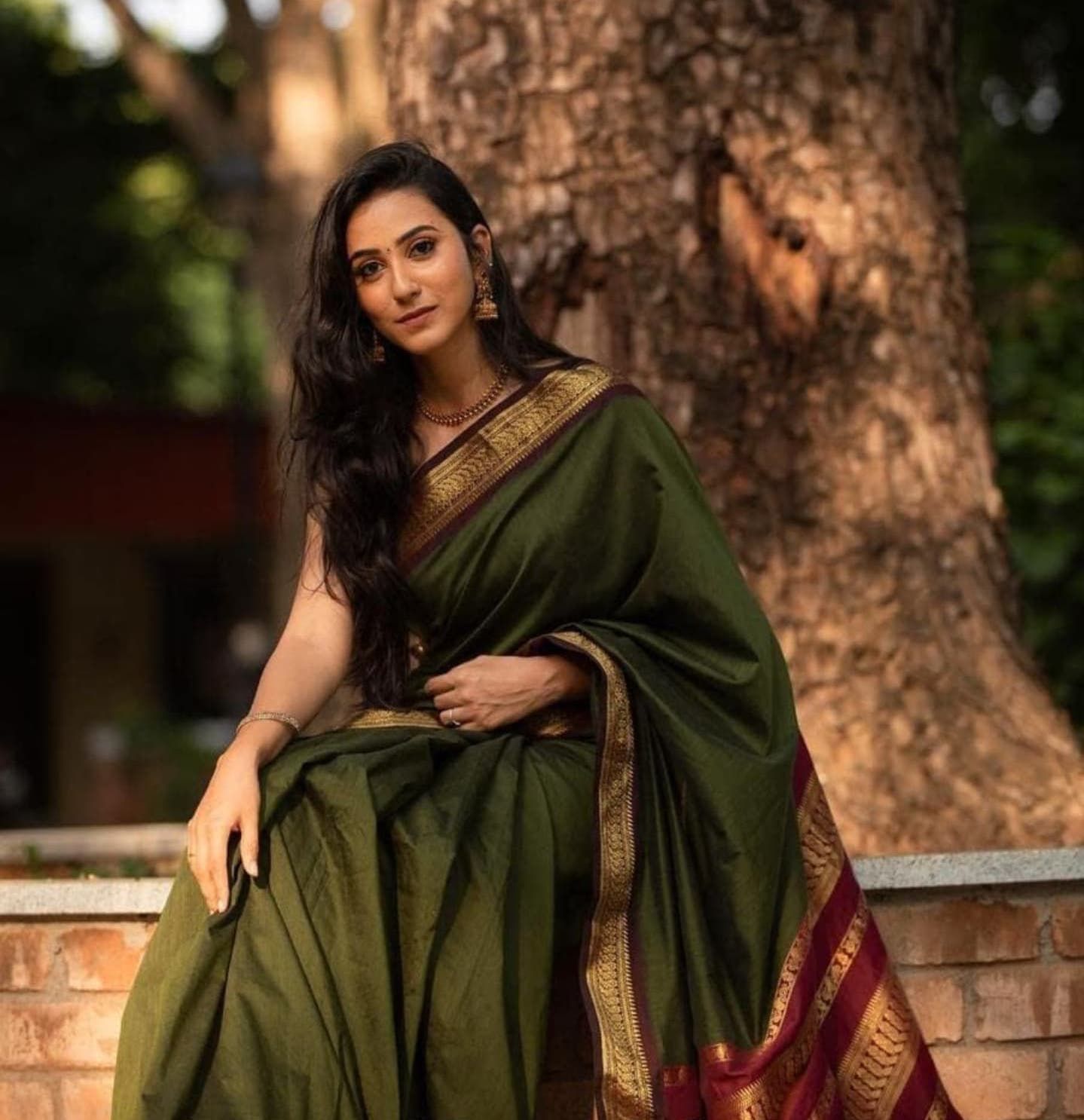 Dark green silk saree with gold border photographed against a tree trunk backdrop.