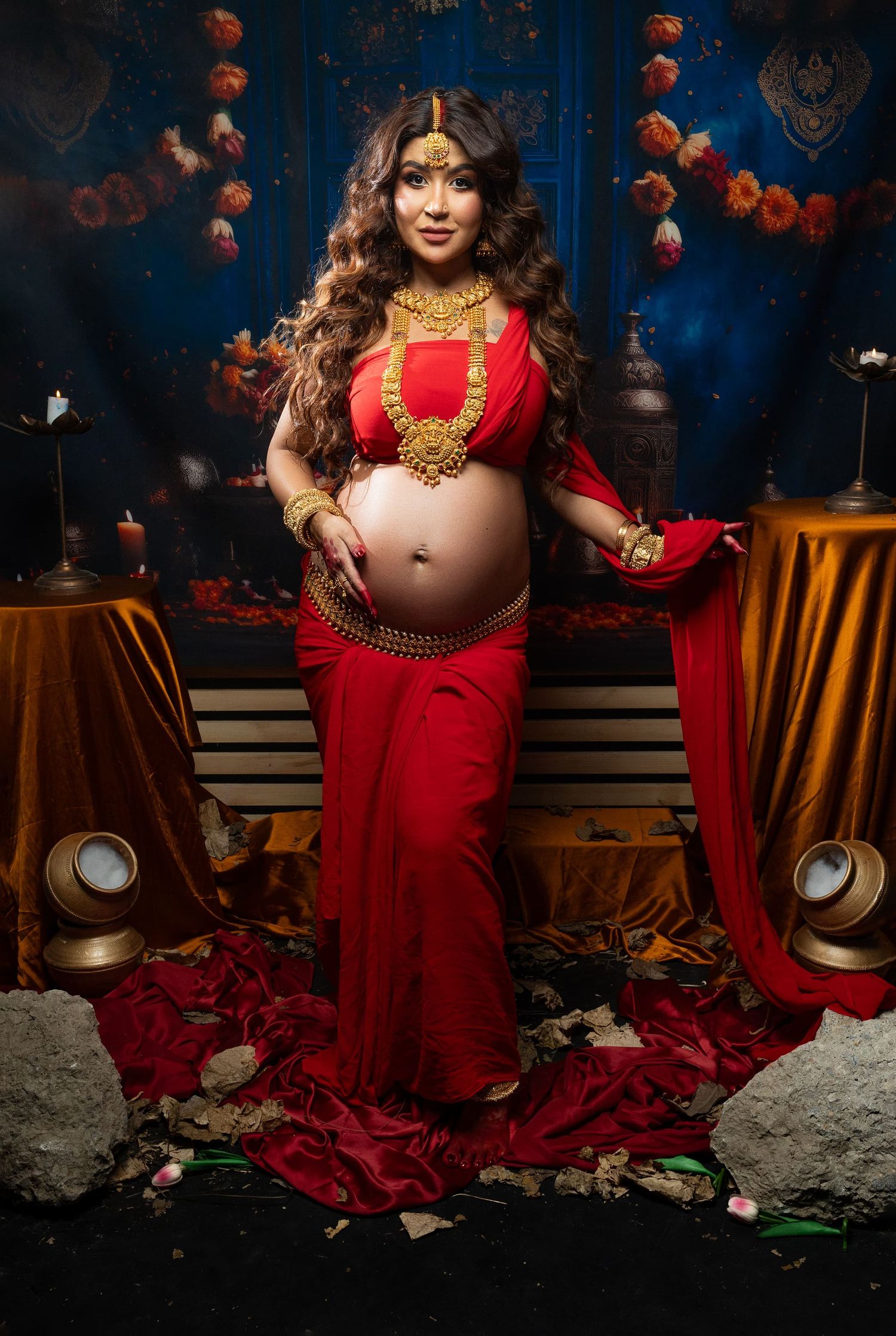 Maternity photoshoot in a vibrant red sari with gold jewelry against a blue backdrop with lanterns and rose petals.