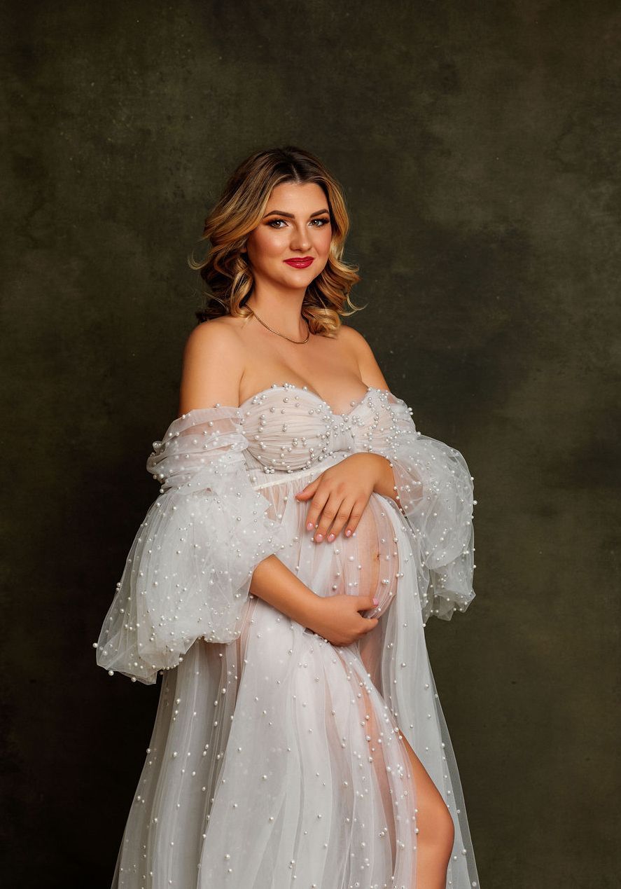 Romantic off-shoulder white lace gown with pearl details against dark backdrop for maternity photoshoot.