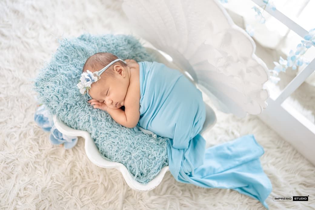 Newborn baby girl sleeping in white shell-shaped bed, wrapped in blue swaddle with floral headband.