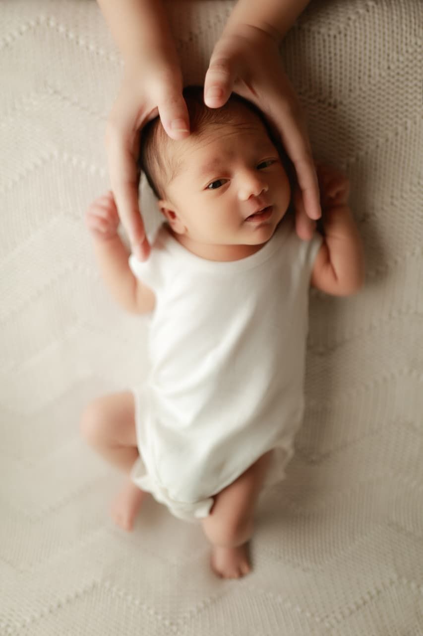 Newborn baby in white onesie lying on blanket with parent hands gently cradling head from above.