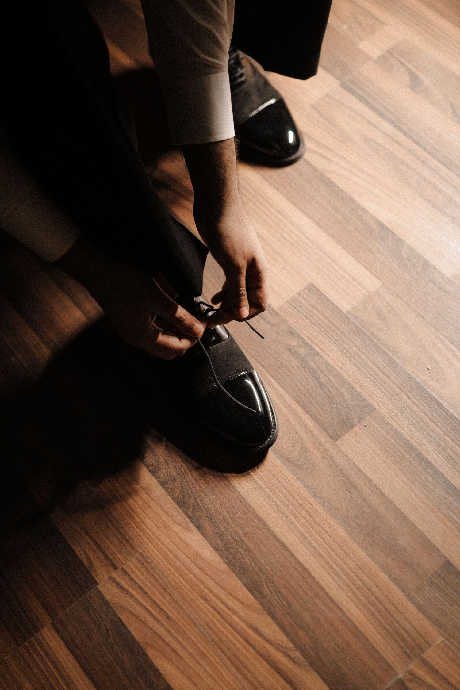 Man tying laces on black dress shoes on wooden floor.