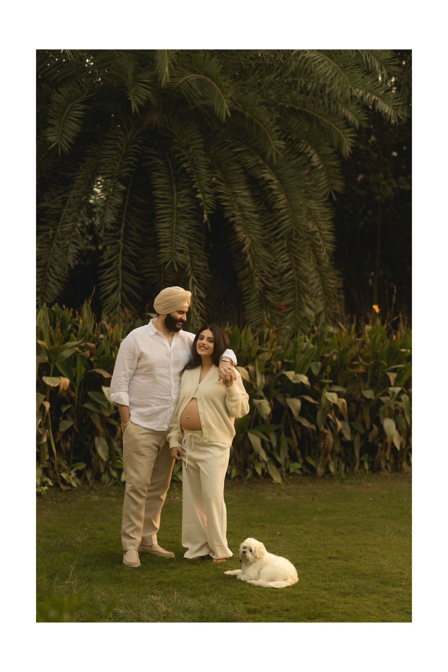 A couple in white outfits poses together in a garden setting with lush greenery and a white dog nearby.