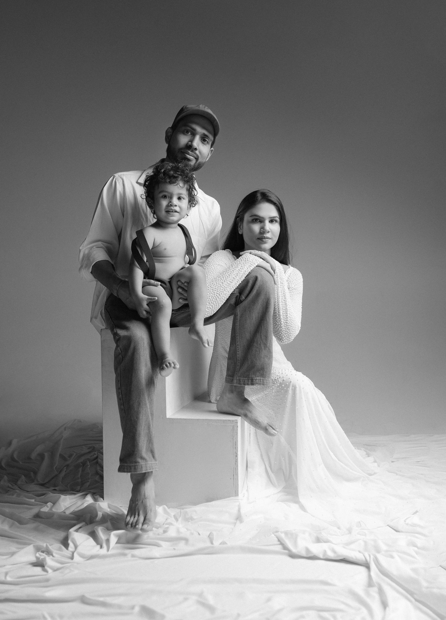A black and white studio portrait of a family sitting together on a white cube against a light background.