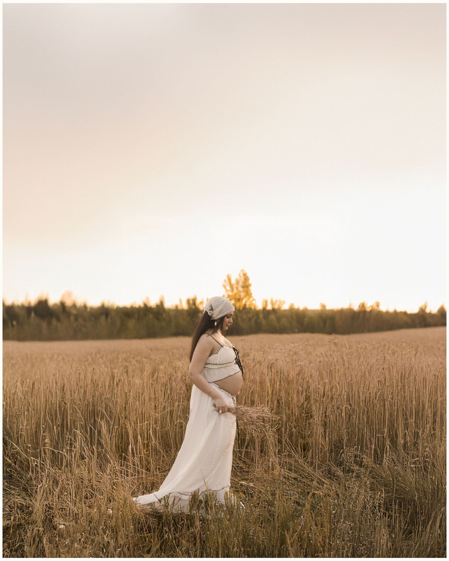 Pregnant woman in white dress and hat stands in golden field at sunset, cradling her baby bump.