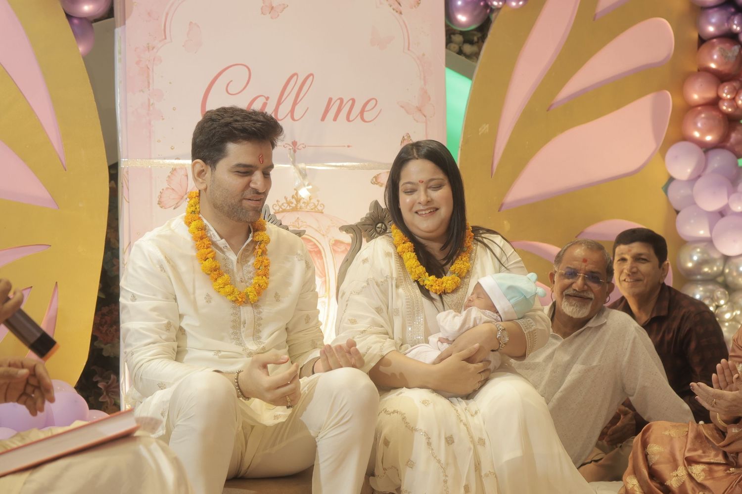 Family celebrates a baby naming ceremony wearing marigold garlands, with a woman holding a newborn beside pink butterfly and balloon decor.