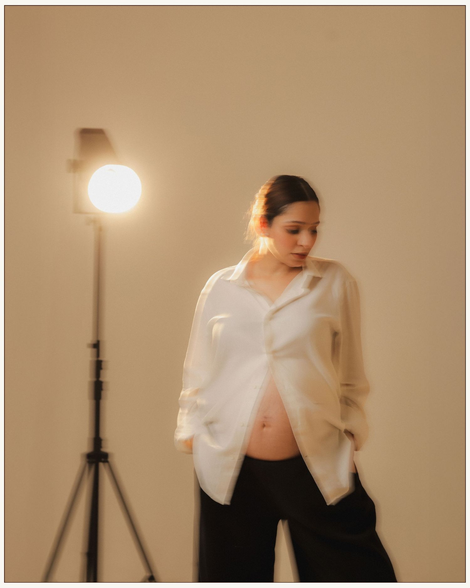 Pregnant woman in white shirt and black pants poses in studio with warm lighting on tripod stand.