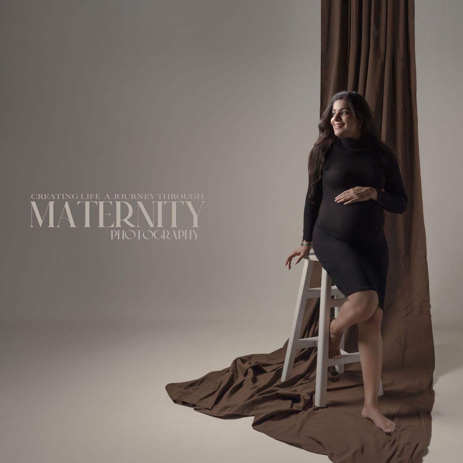Studio maternity photo with brown drapery and minimalist chair prop in muted tones.