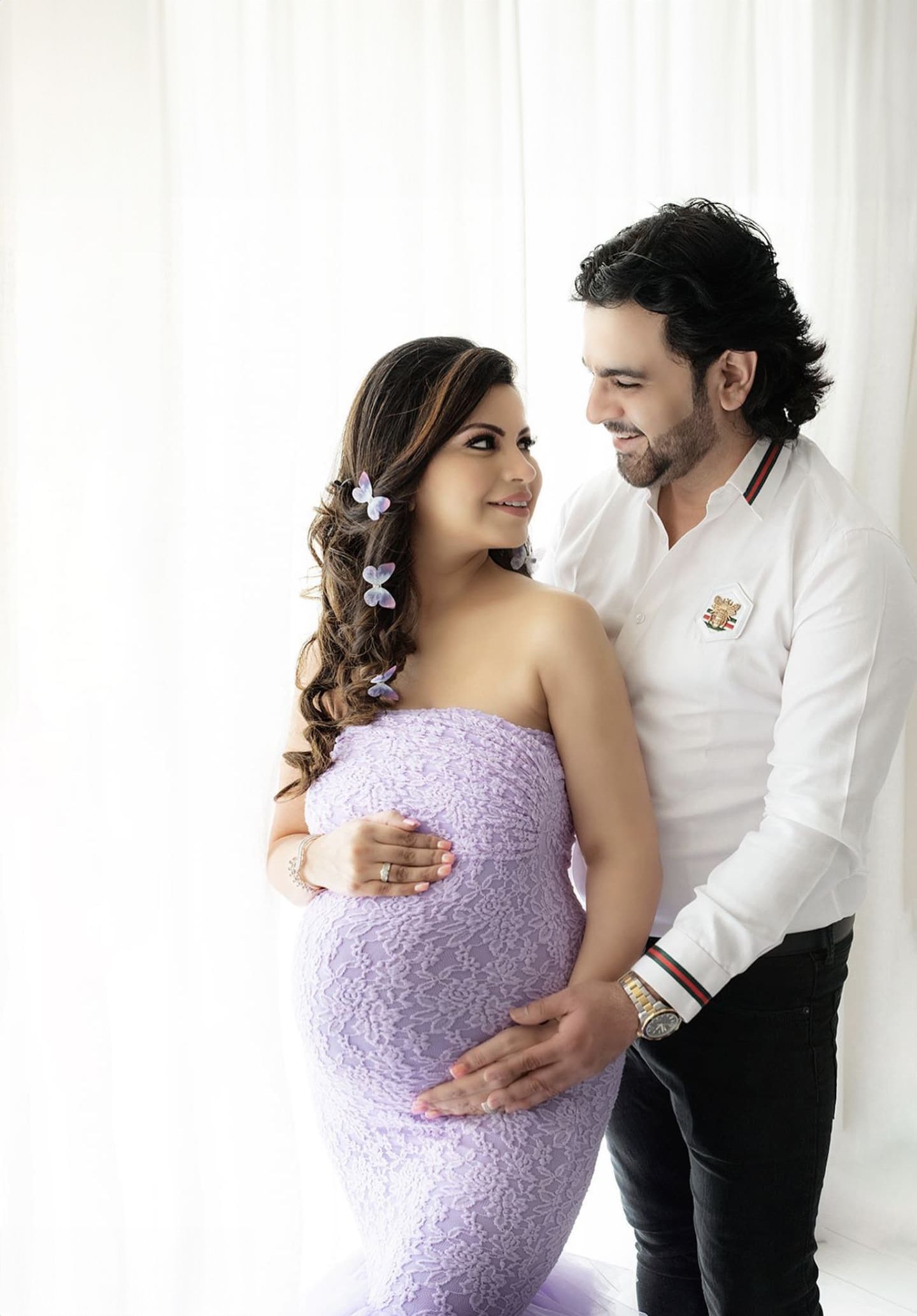 Intimate maternity portrait with couple in lavender lace gown and white shirt against bright background.