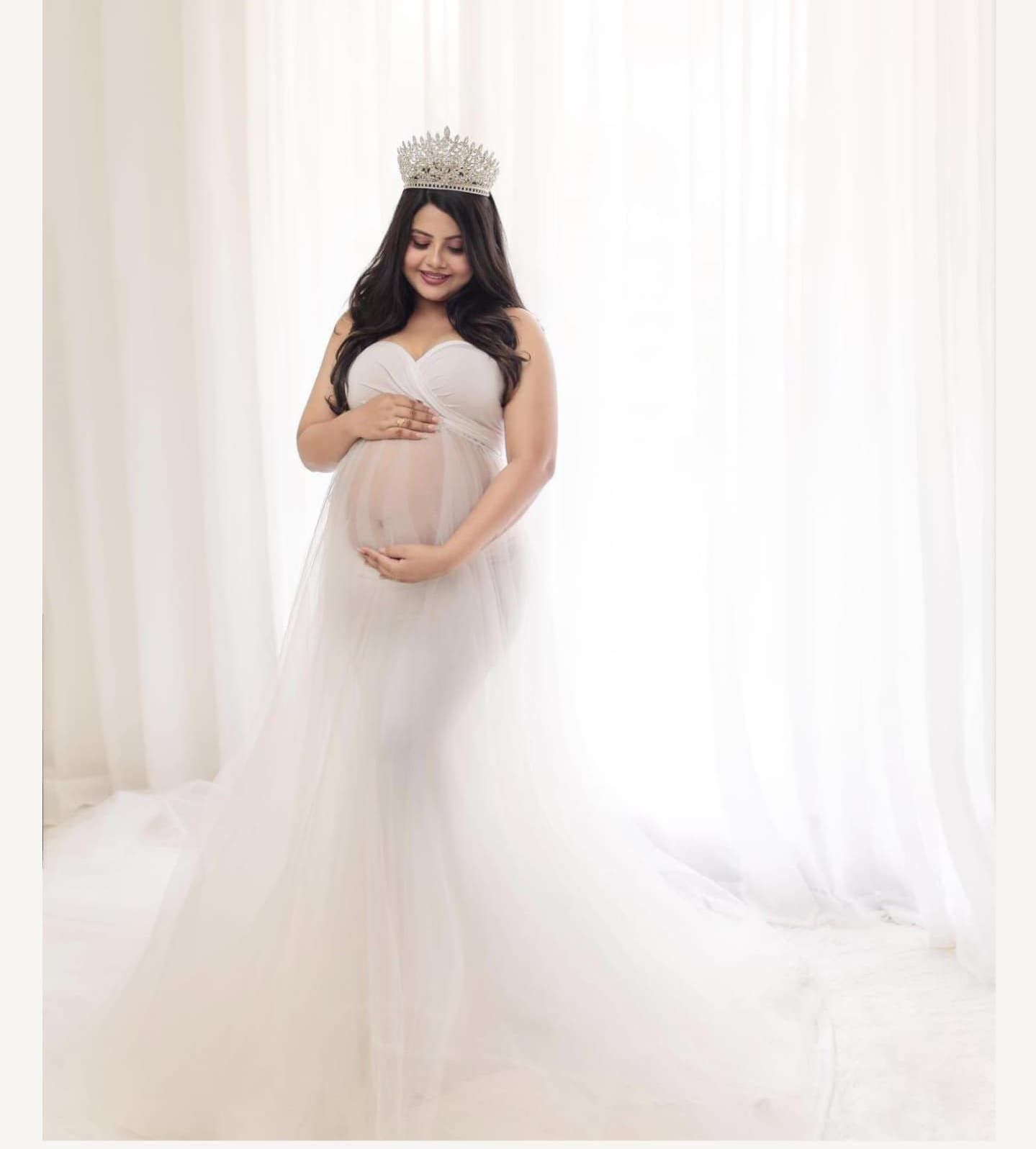 A maternity photoshoot featuring an elegant white gown and crown against a bright window backdrop.
