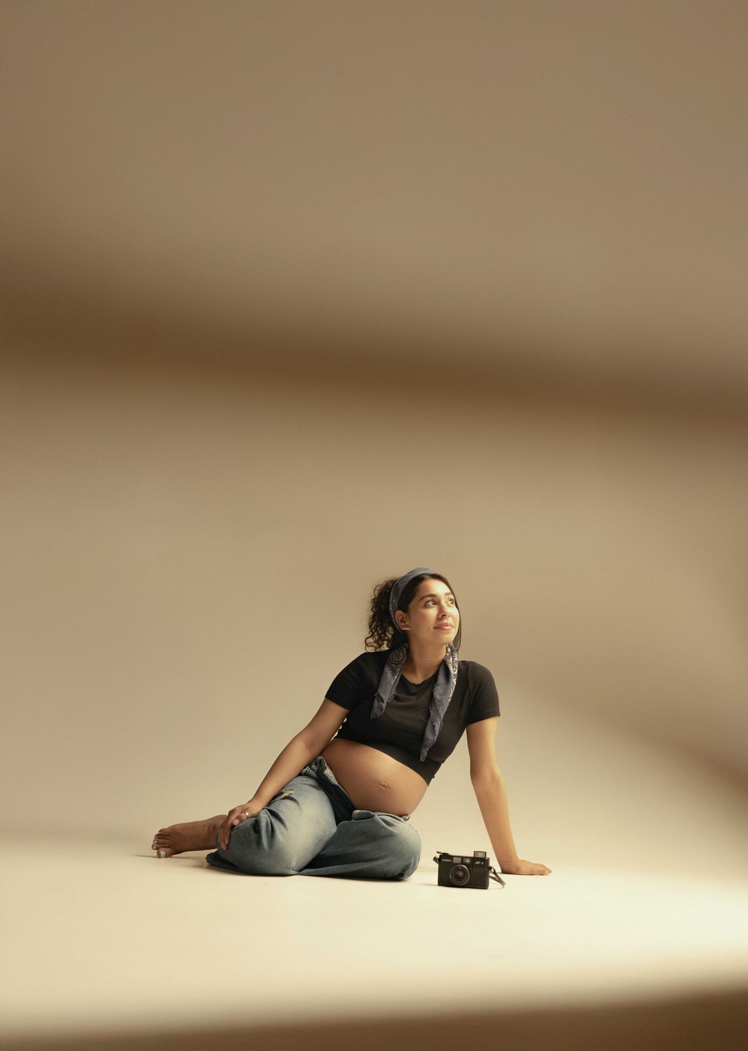 Pregnant woman in black crop top and jeans sitting on floor, gazing upward in a minimalist beige studio.