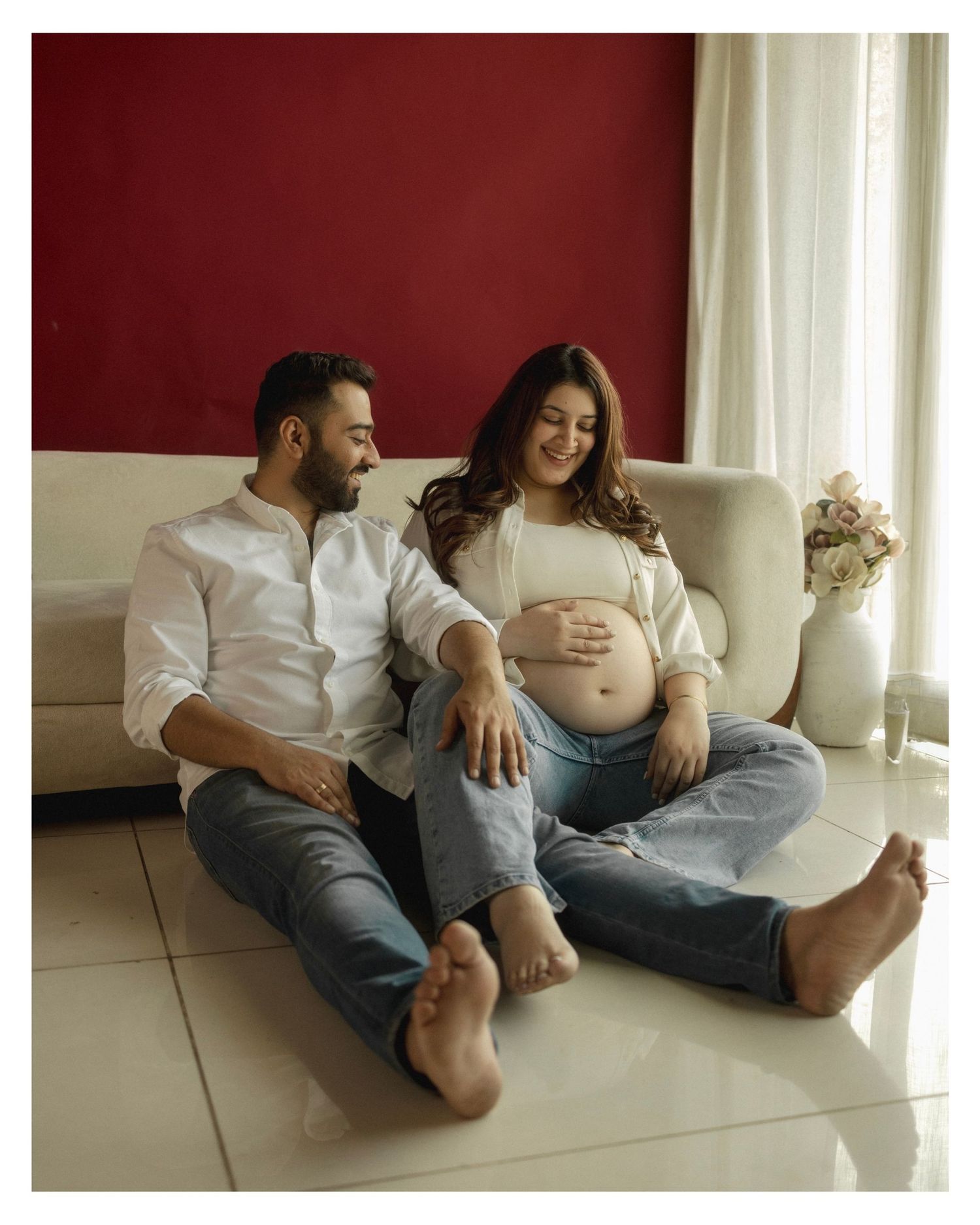 Couple sitting on floor, pregnant woman cradling baby bump, man smiling beside her, red wall background.