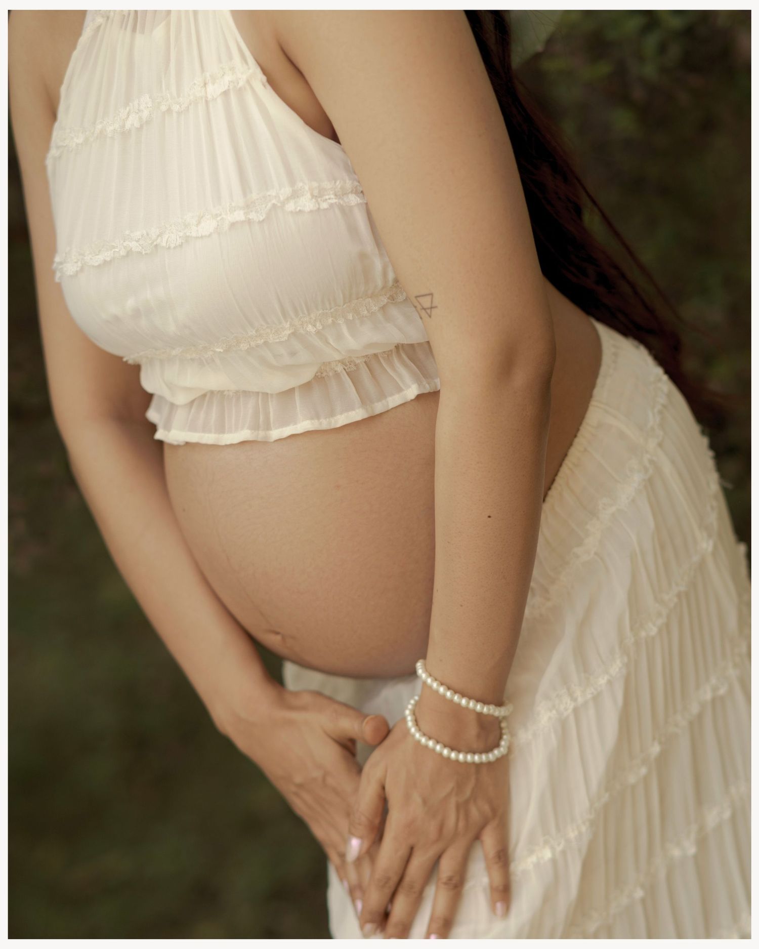 Pregnant woman in white ruffled crop top and skirt, cradling baby bump, wearing pearl bracelet, outdoor maternity photo.