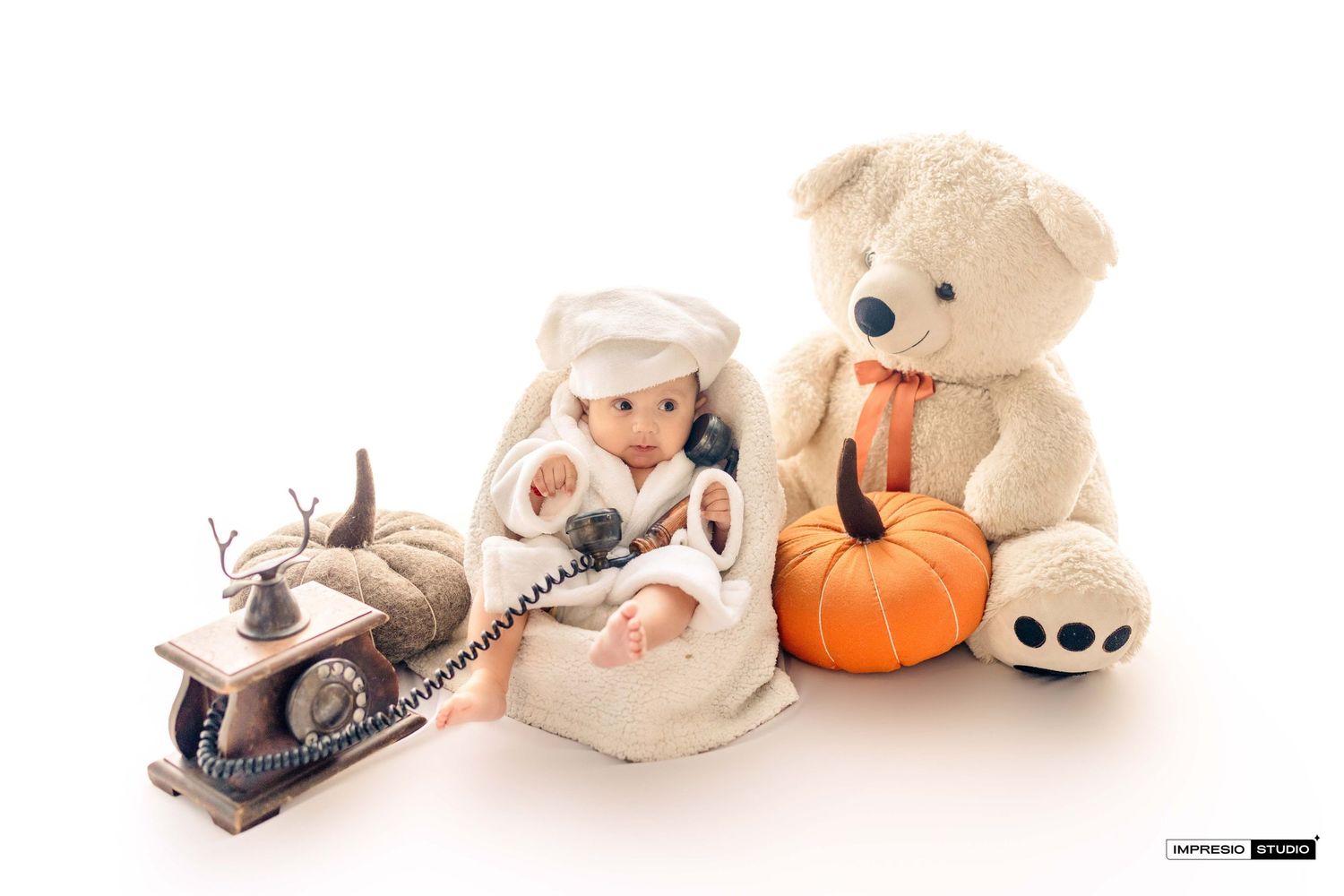 Baby in white costume sits with vintage phone and large teddy bear beside orange pumpkin on white background.