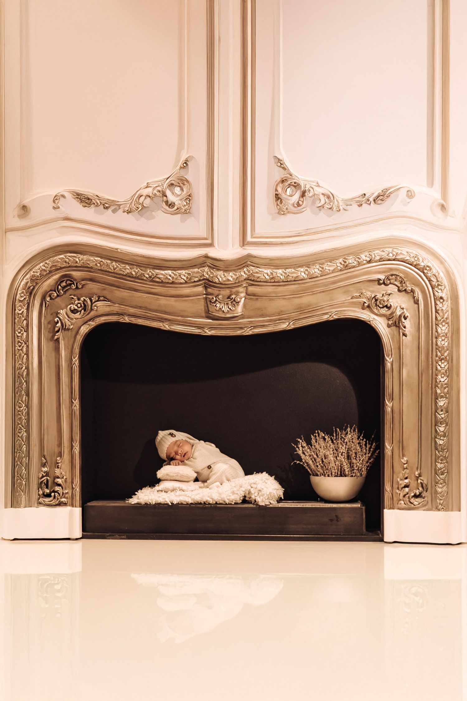 Newborn baby sleeping peacefully inside an ornate gold fireplace mantel with white fluffy blanket and dried lavender.