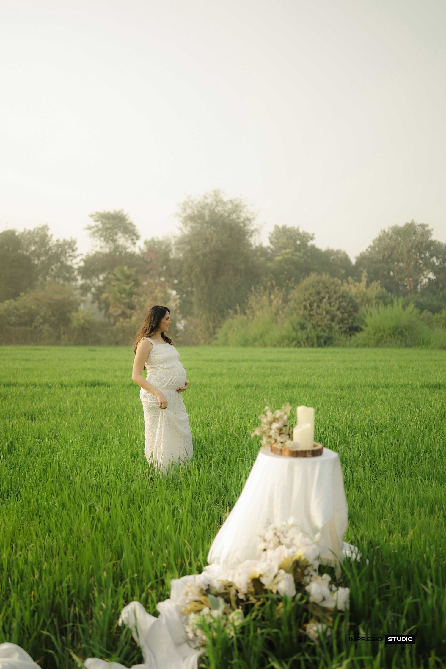 Pregnant woman in white dress stands in lush green field beside a decorated table with flowers and cake.