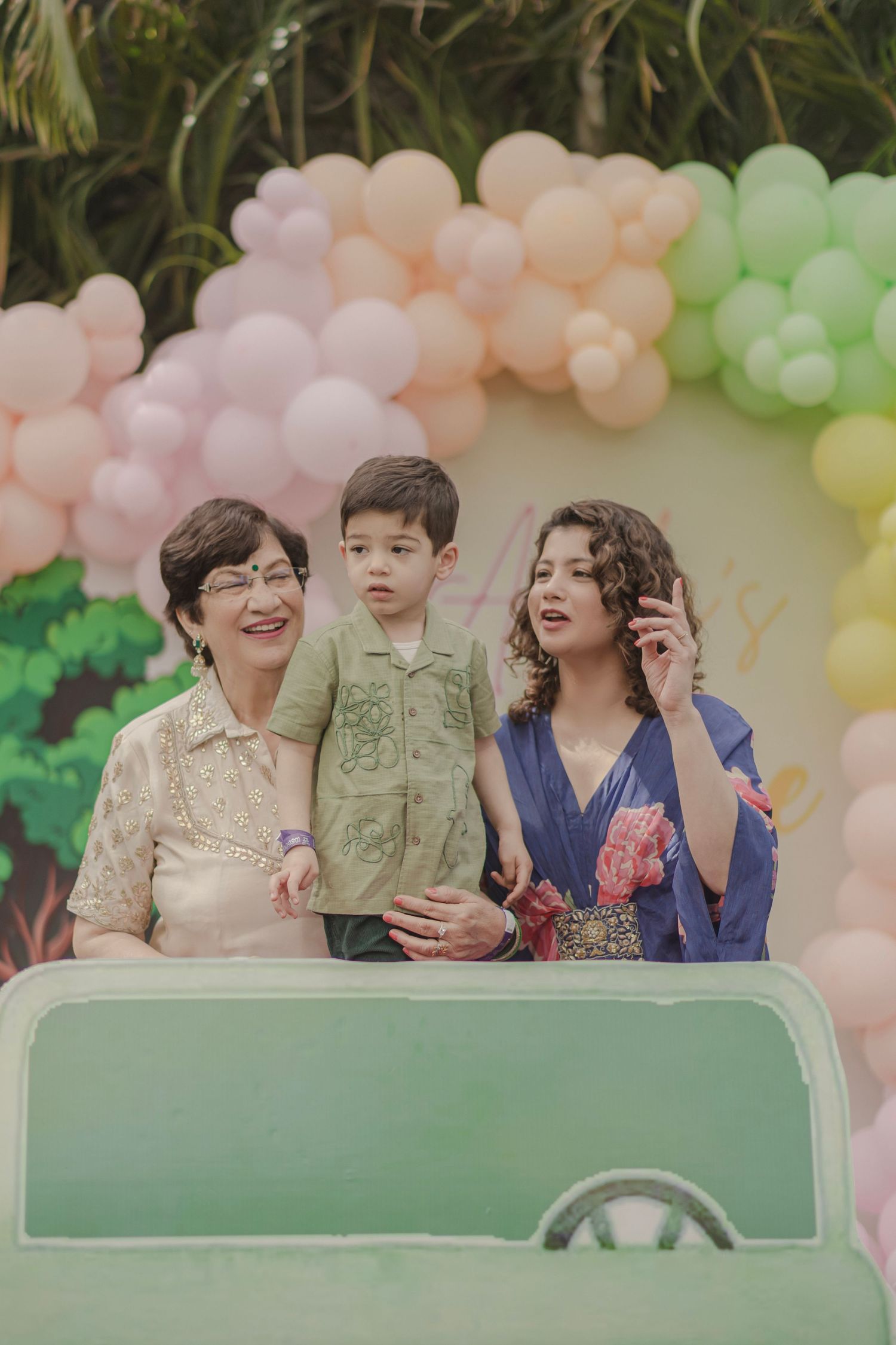 Three generations pose at a colorful balloon-decorated birthday party outdoors.
