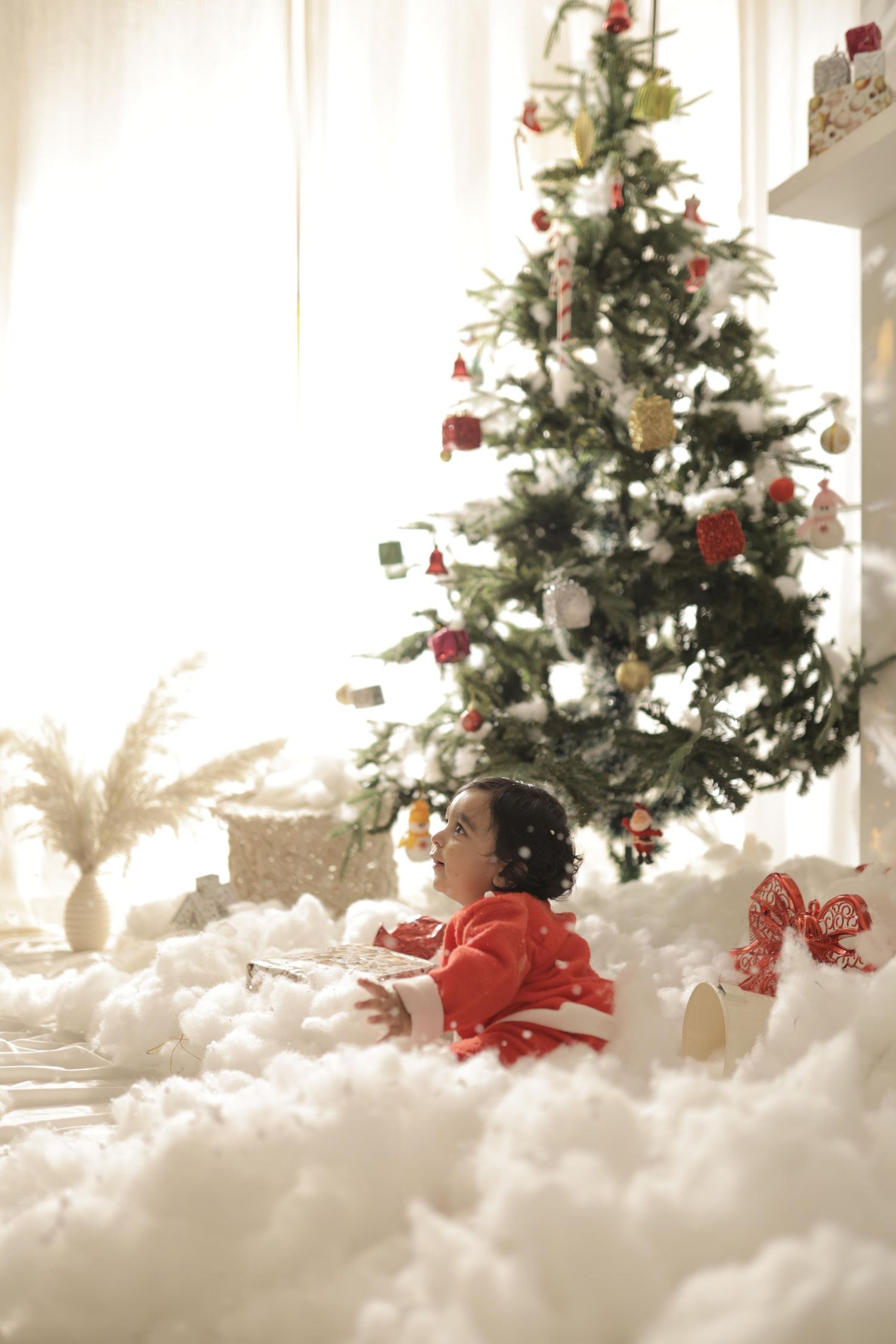 Child in red outfit sitting among fluffy white snow surrounded by Christmas gifts near a decorated holiday tree indoors.