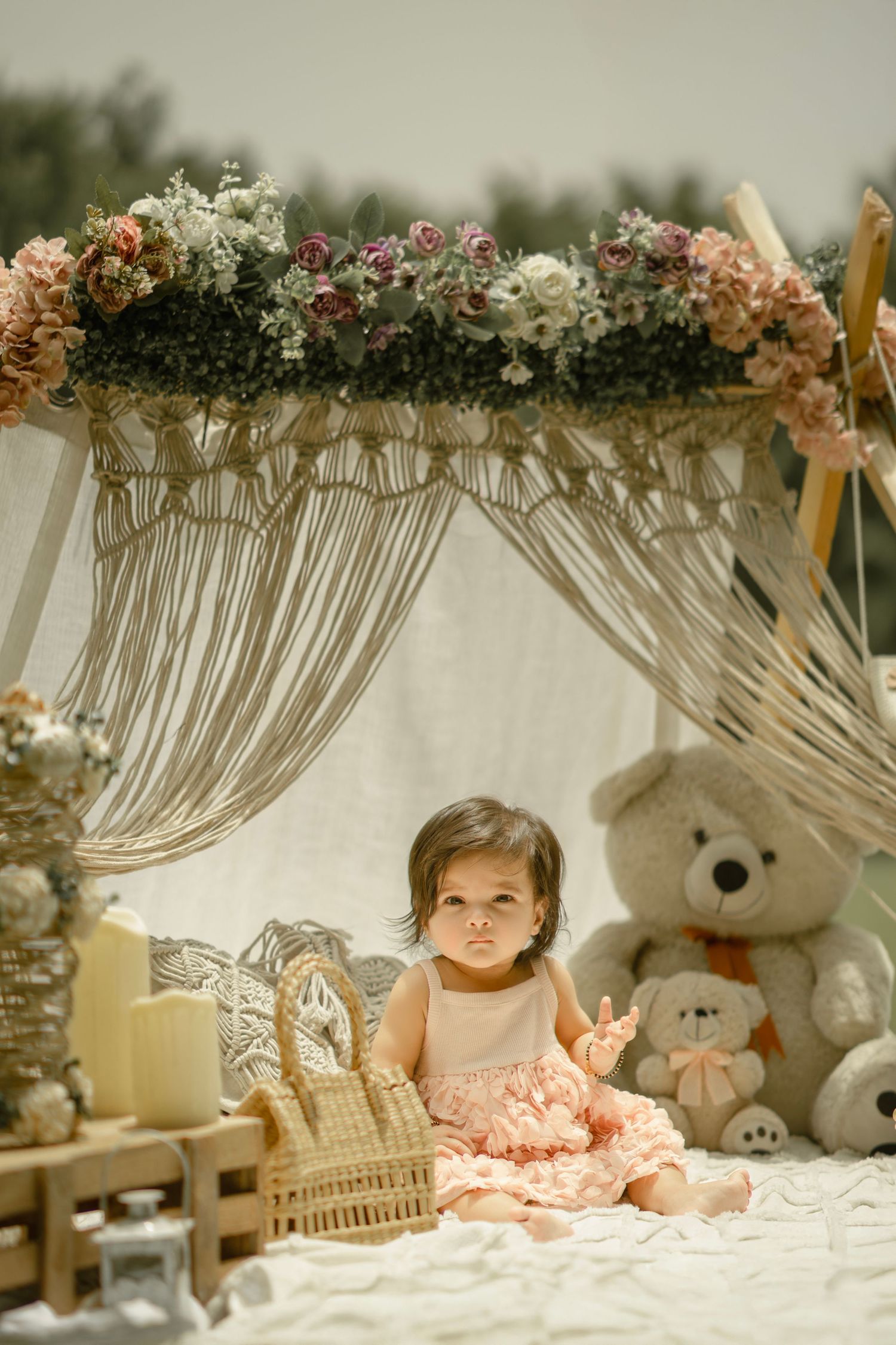 Toddler girl in pink dress sitting under floral macrame canopy with teddy bears and wicker basket outdoors.
