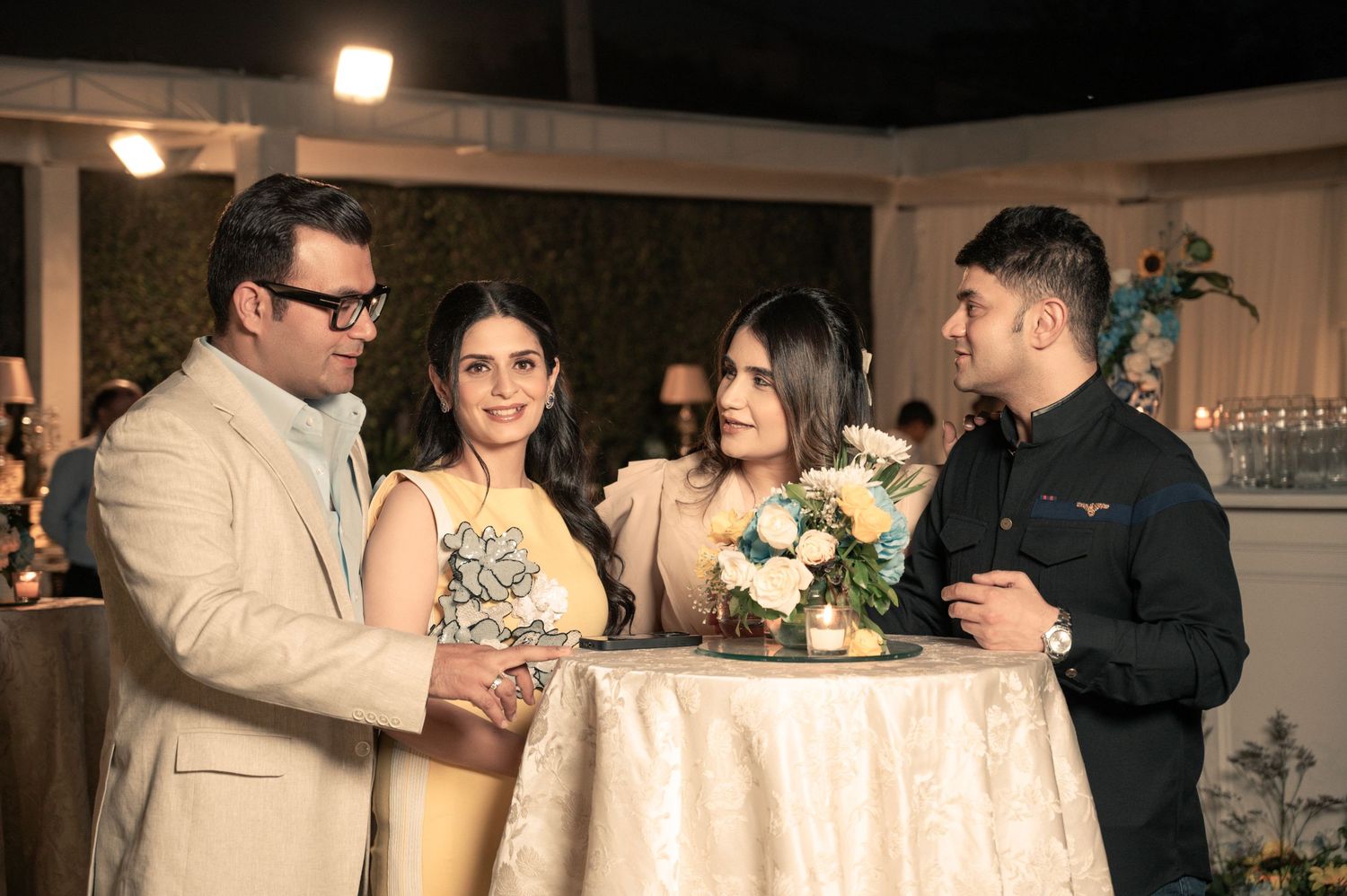 Four elegantly dressed guests socializing around a floral centerpiece table at an indoor evening celebration.