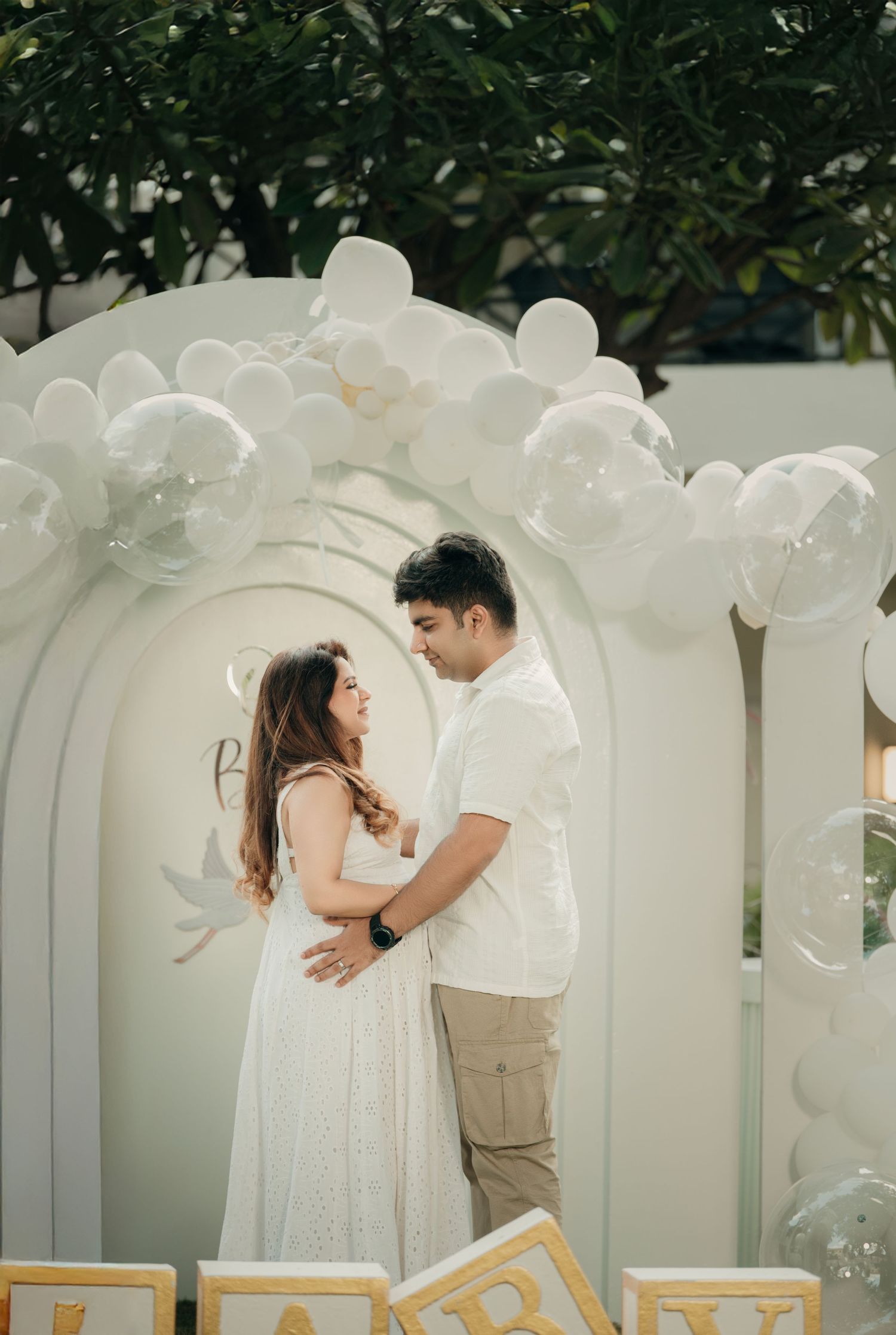 Couple embracing under white balloon arch at elegant outdoor maternity celebration with decorative letter blocks.