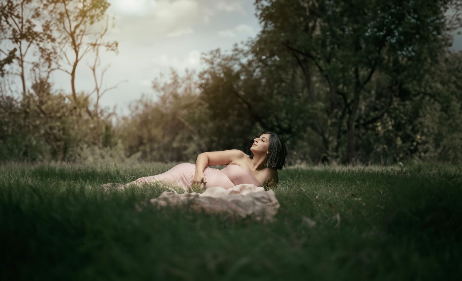 A pregnant woman in a pink dress reclines gracefully on lush green grass surrounded by trees.