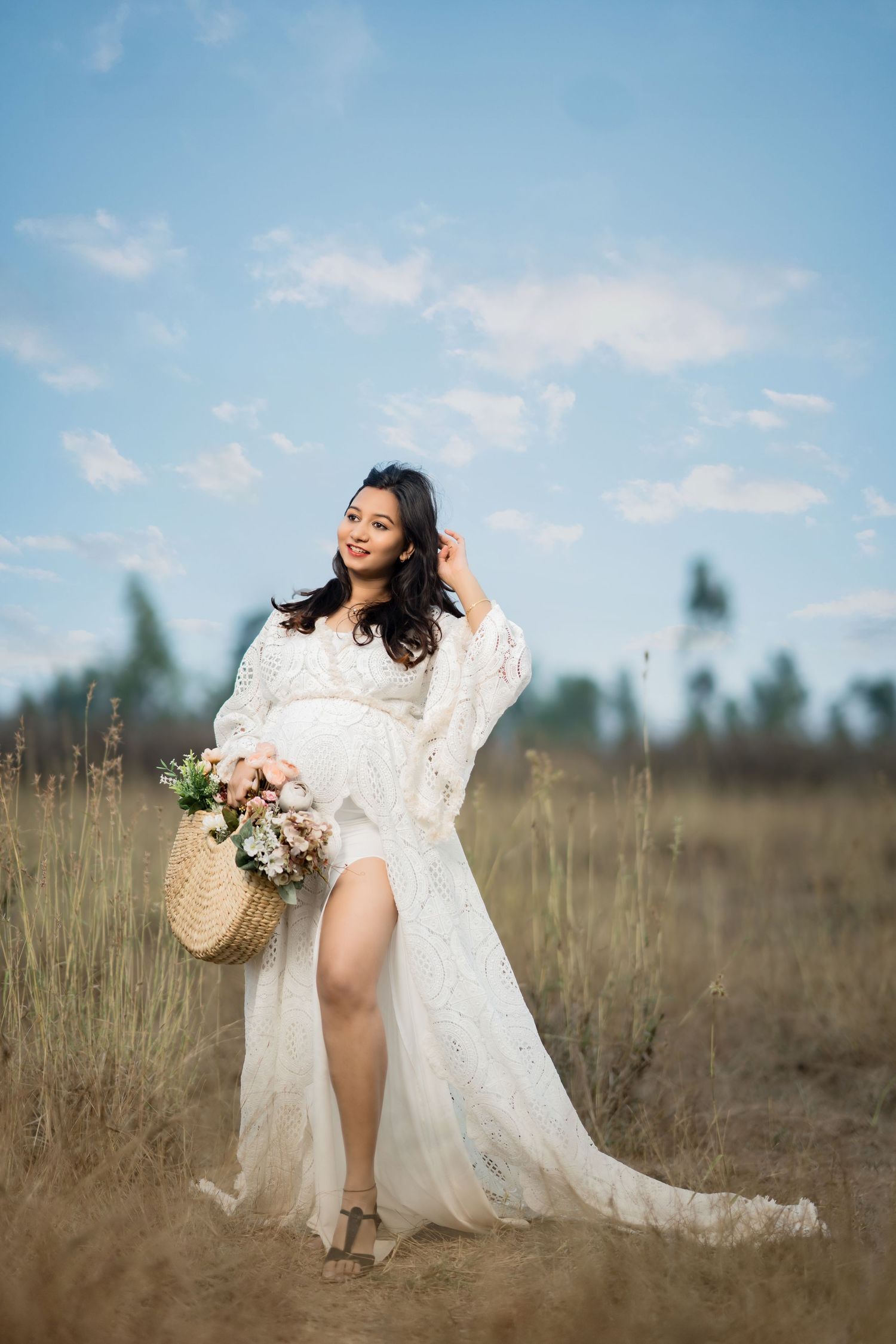 Pregnant woman in white lace dress holding flower basket in field.