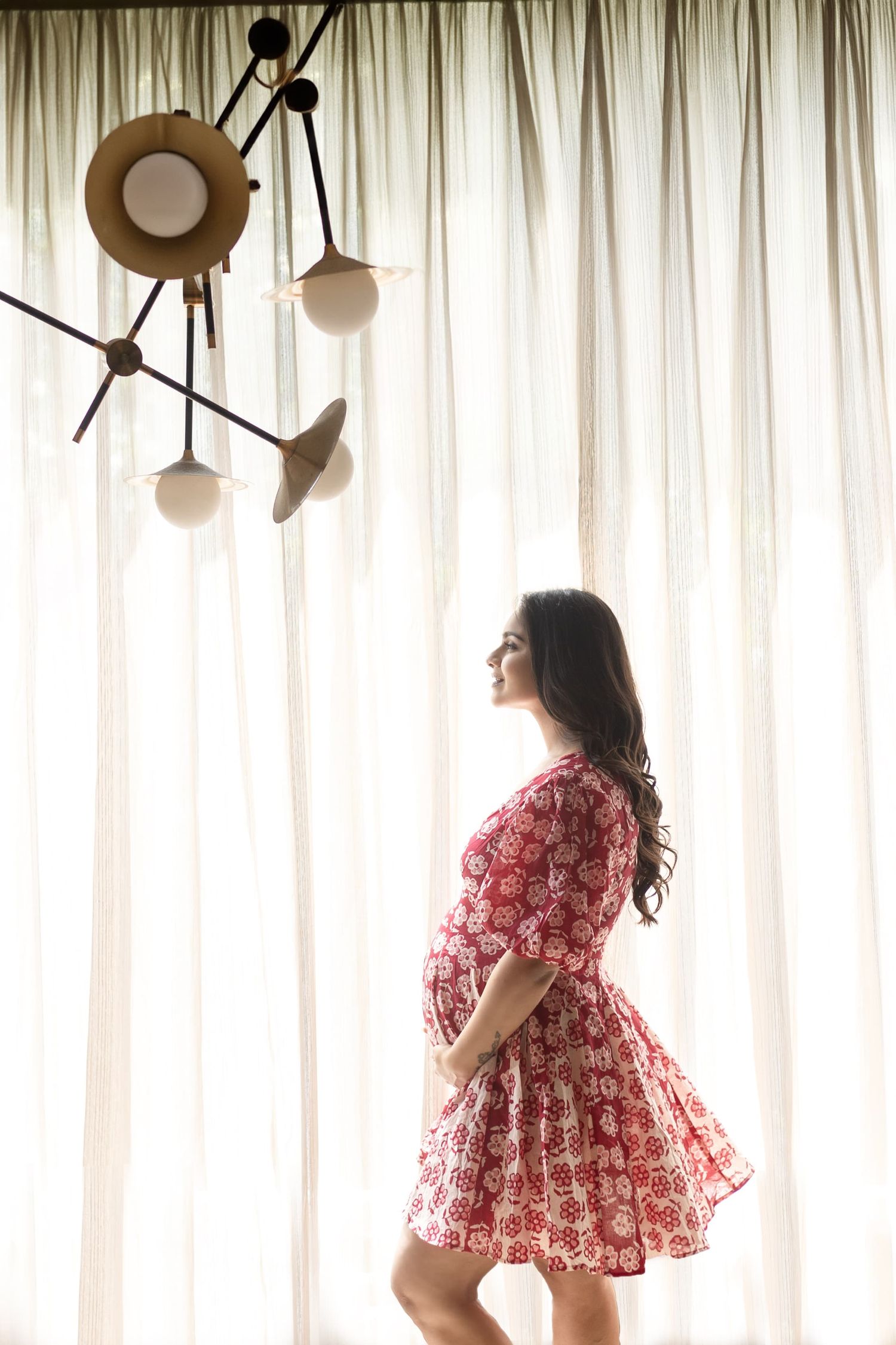Pregnant woman in red floral dress standing by sheer curtains, cradling baby bump, with modern chandelier overhead.