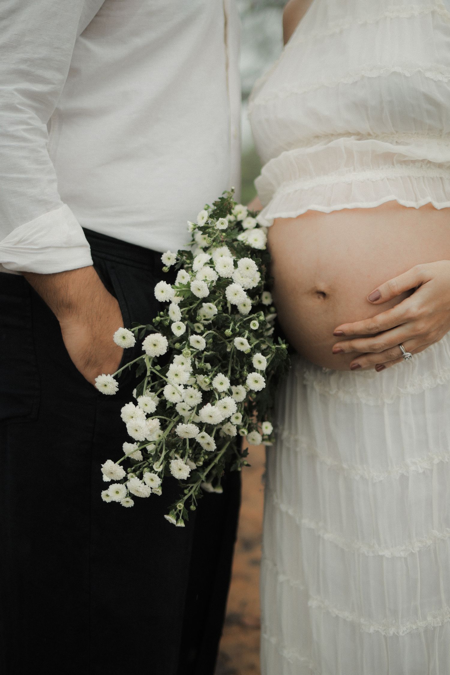Close-up of pregnant belly in white dress with person holding delicate white baby's breath flowers against dark clothing.