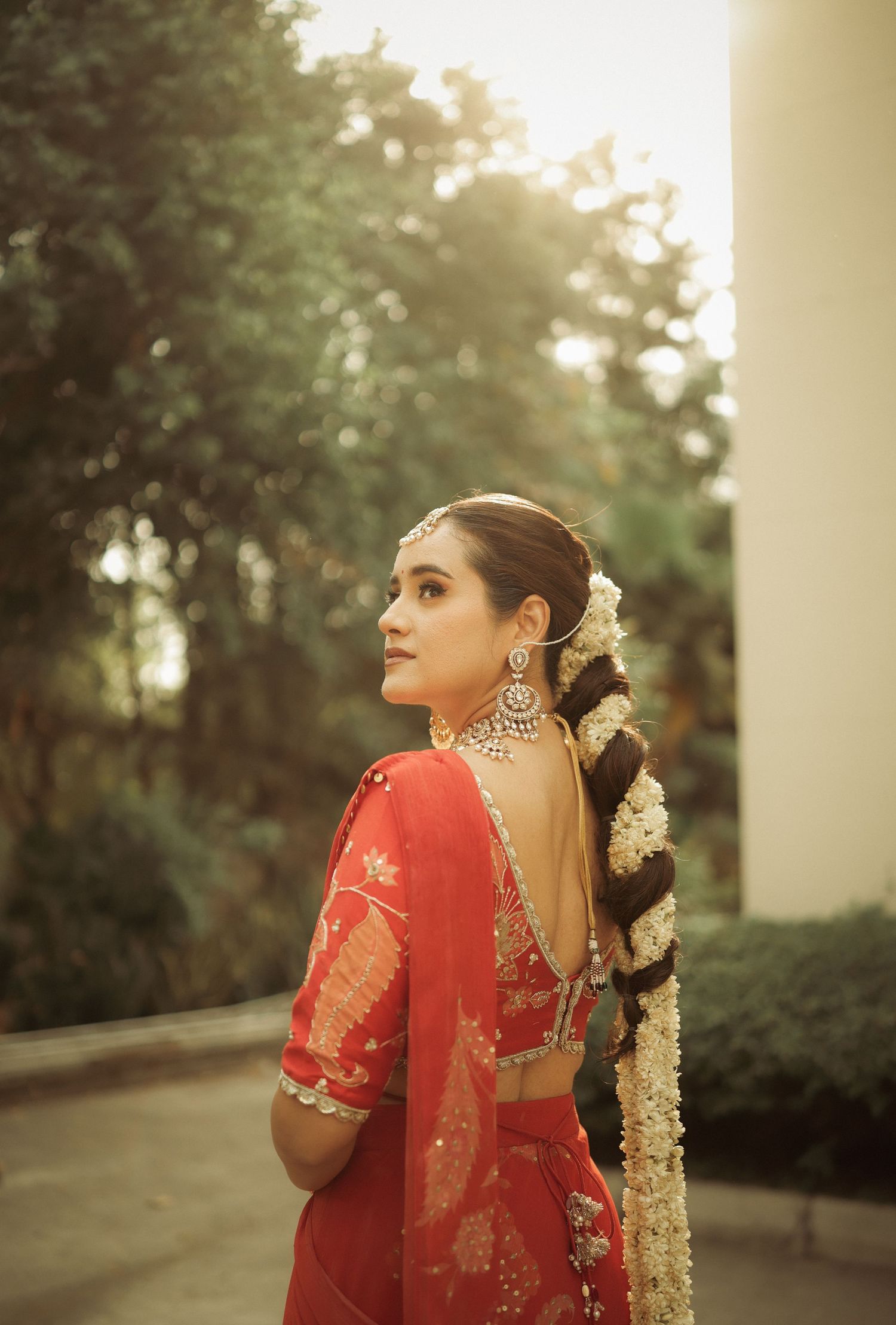Woman in red backless lehenga with floral braid and chandbali earrings poses outdoors in bridal attire.