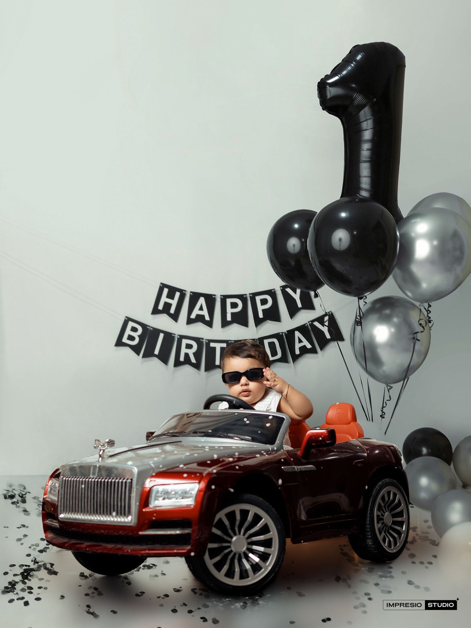First birthday celebration setup with a red toy Rolls Royce car, black and silver balloons, and Happy Birthday banner.