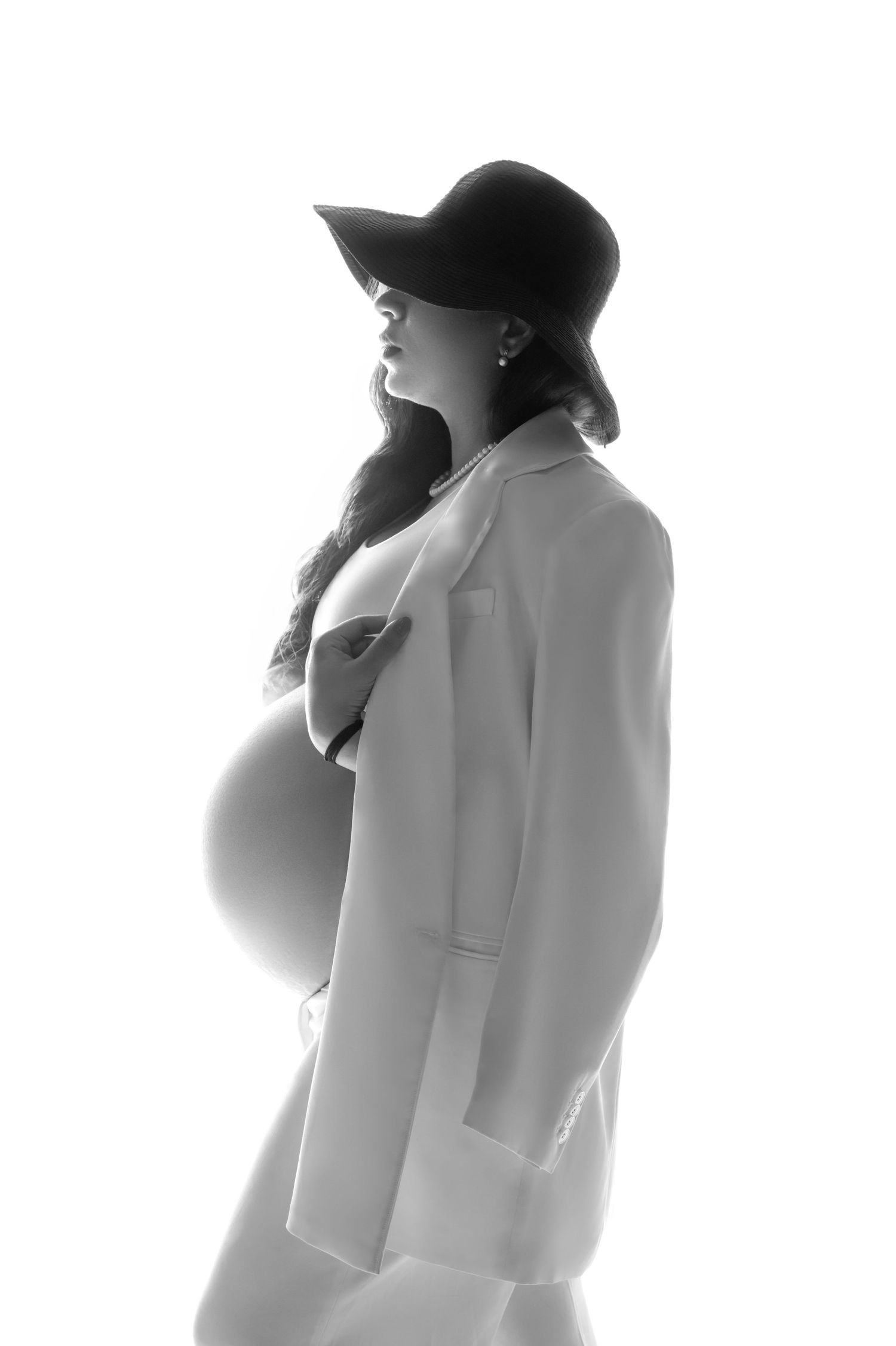 Black and white profile portrait of a pregnant woman in a wide-brim hat and open blazer, showcasing her baby bump.
