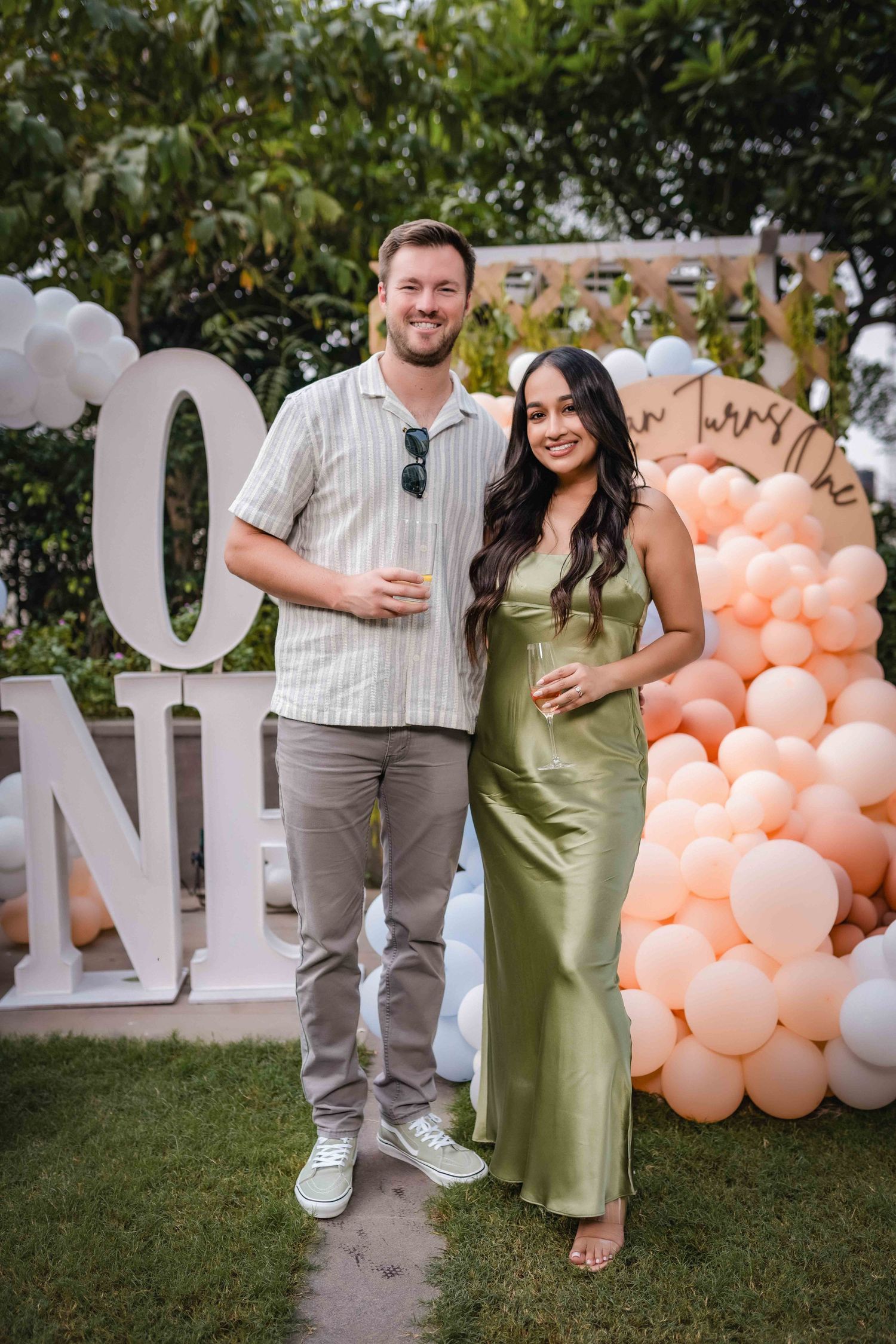 Couple posing together at an outdoor baby shower with peach balloon arch and white letter decorations.