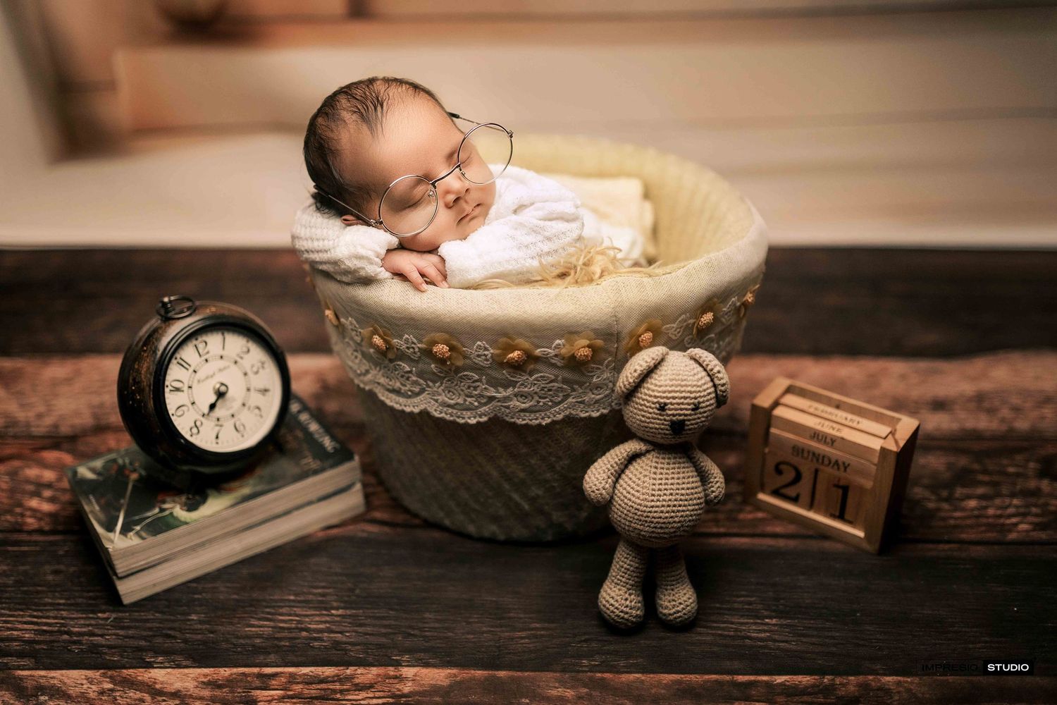 Sleeping newborn in knit basket with teddy bear, clock, and wooden blocks.