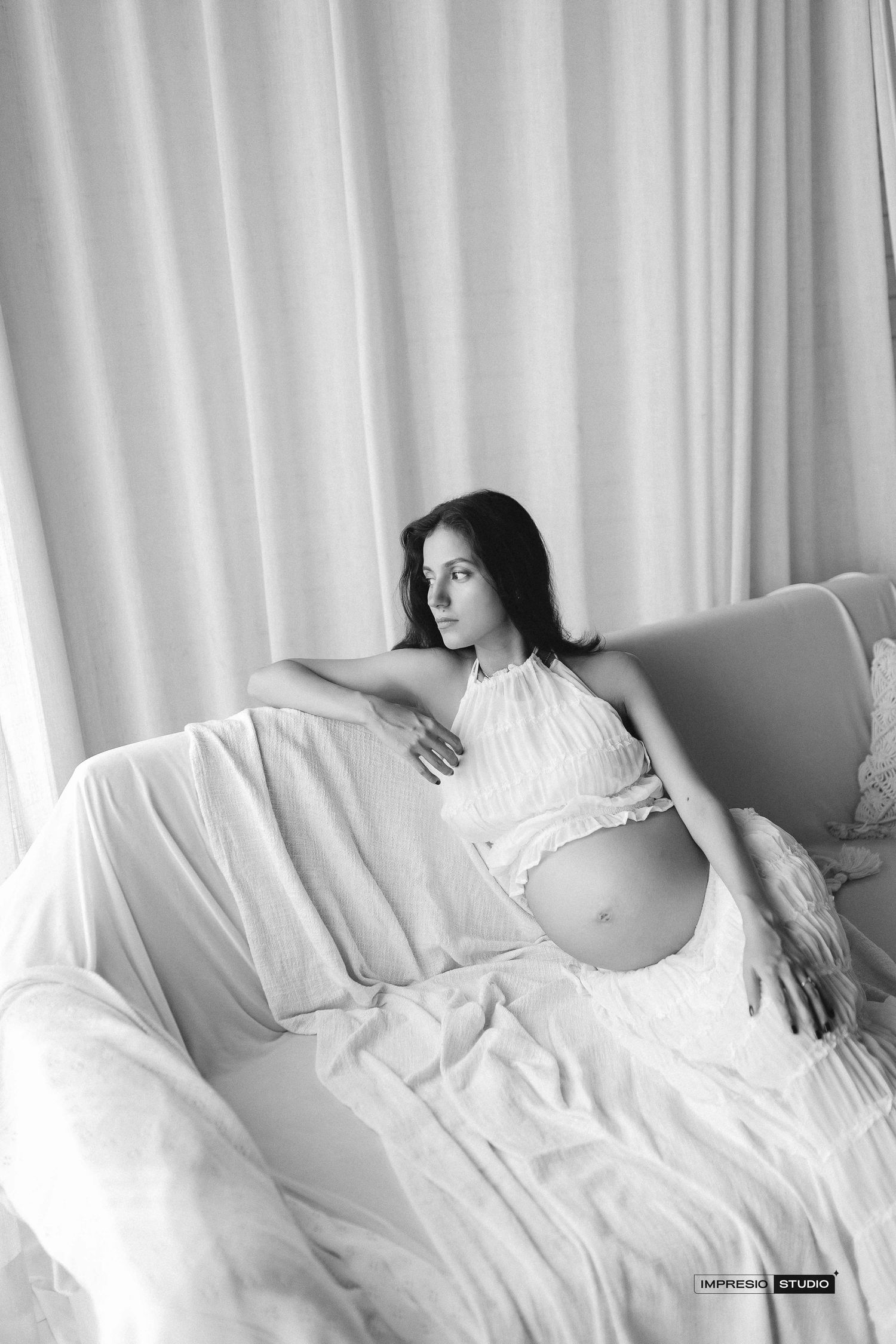 Black and white maternity photo of a pregnant woman in a white halter dress reclining on a sofa near sheer curtains.