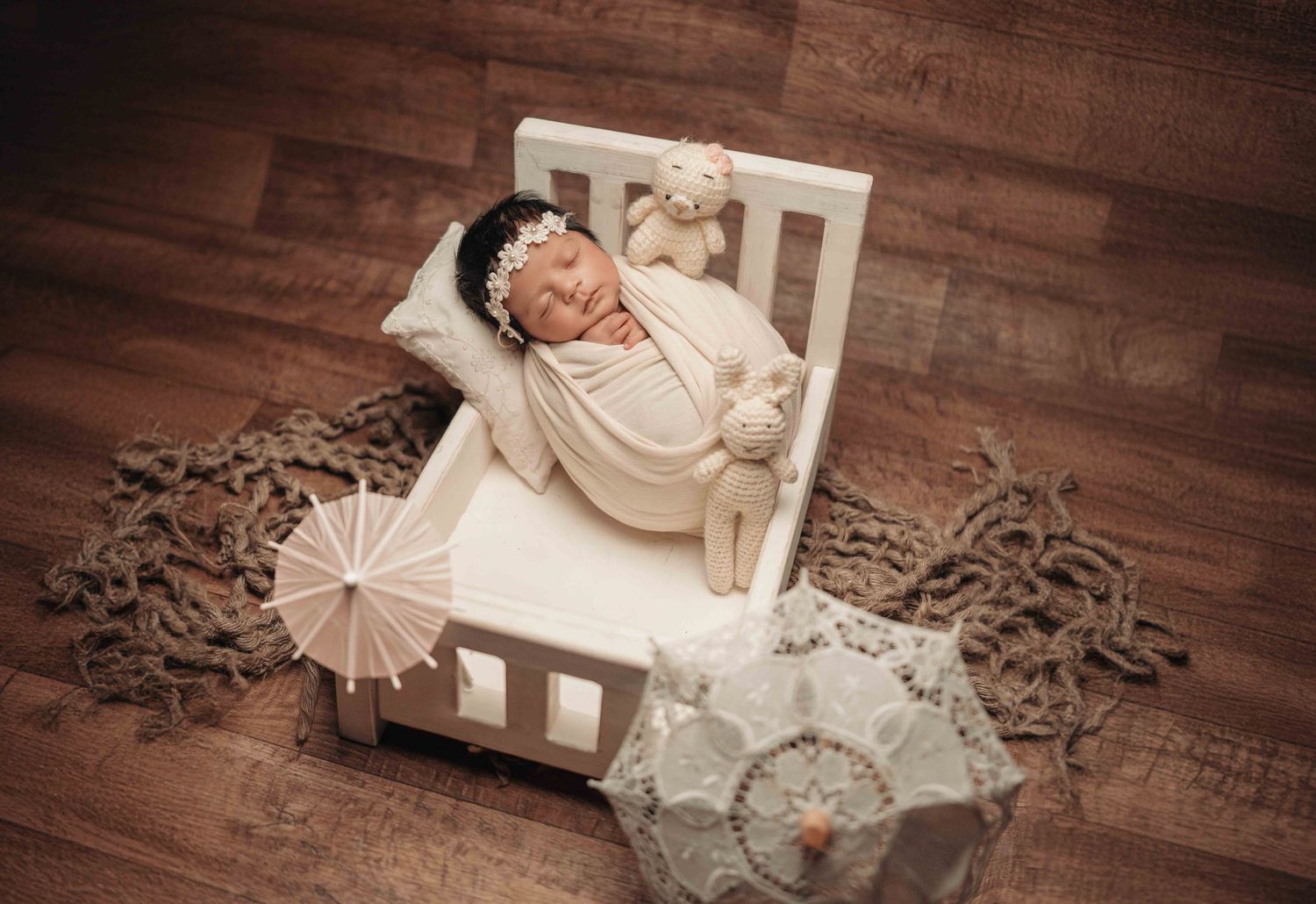 Newborn baby swaddled in cream wrap sleeping peacefully in a tiny white miniature bed with soft toys.