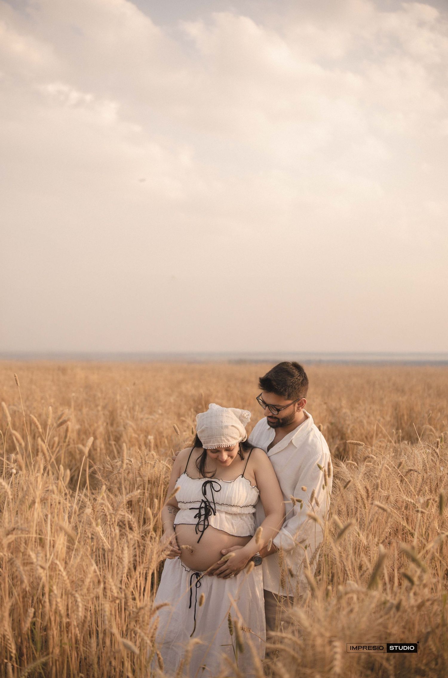 Couple embracing in golden wheat field during maternity photoshoot, woman in white dress showing baby bump at sunset.