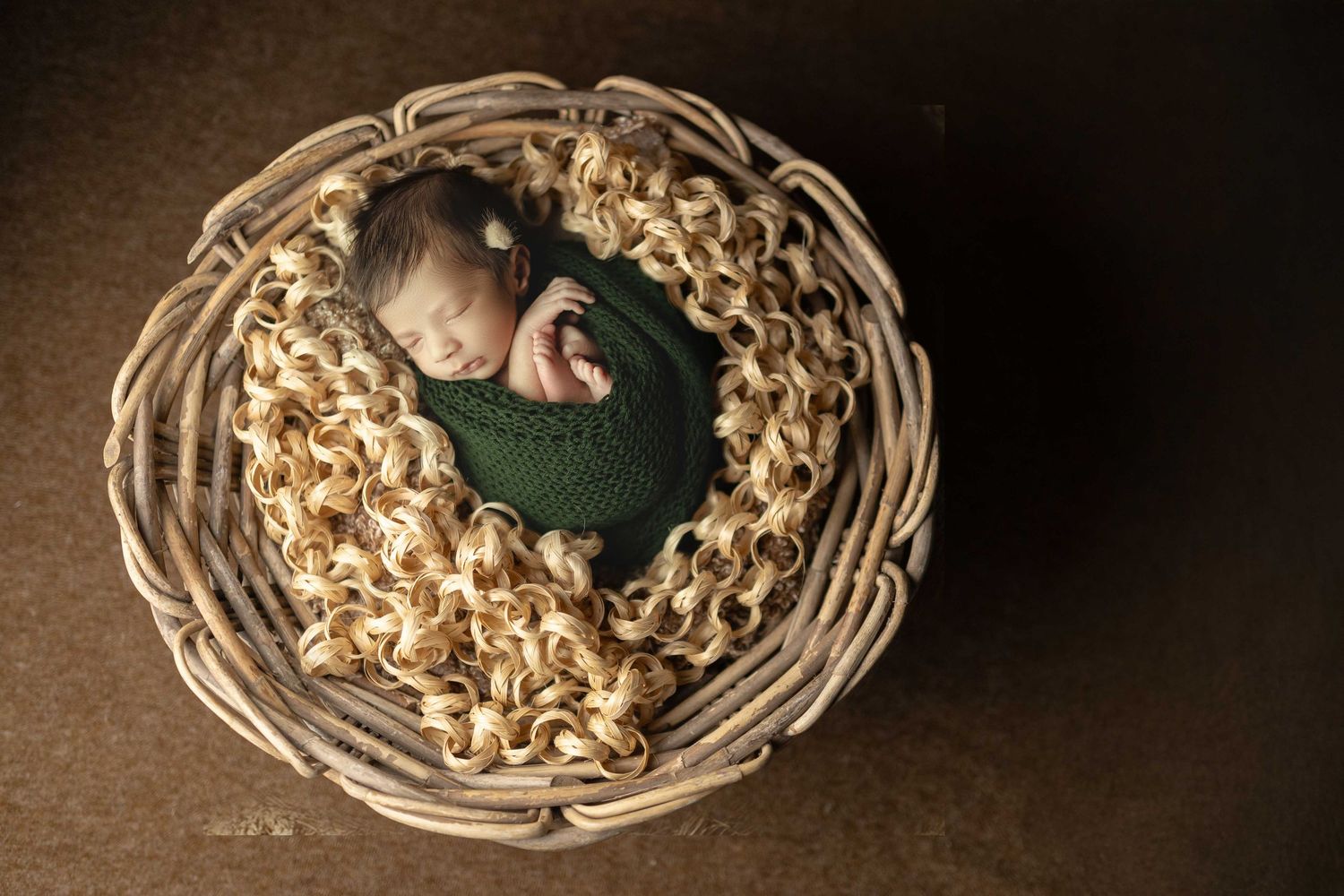 Newborn baby sleeping peacefully in a wicker basket wrapped in a green knit swaddle on a soft textured blanket.